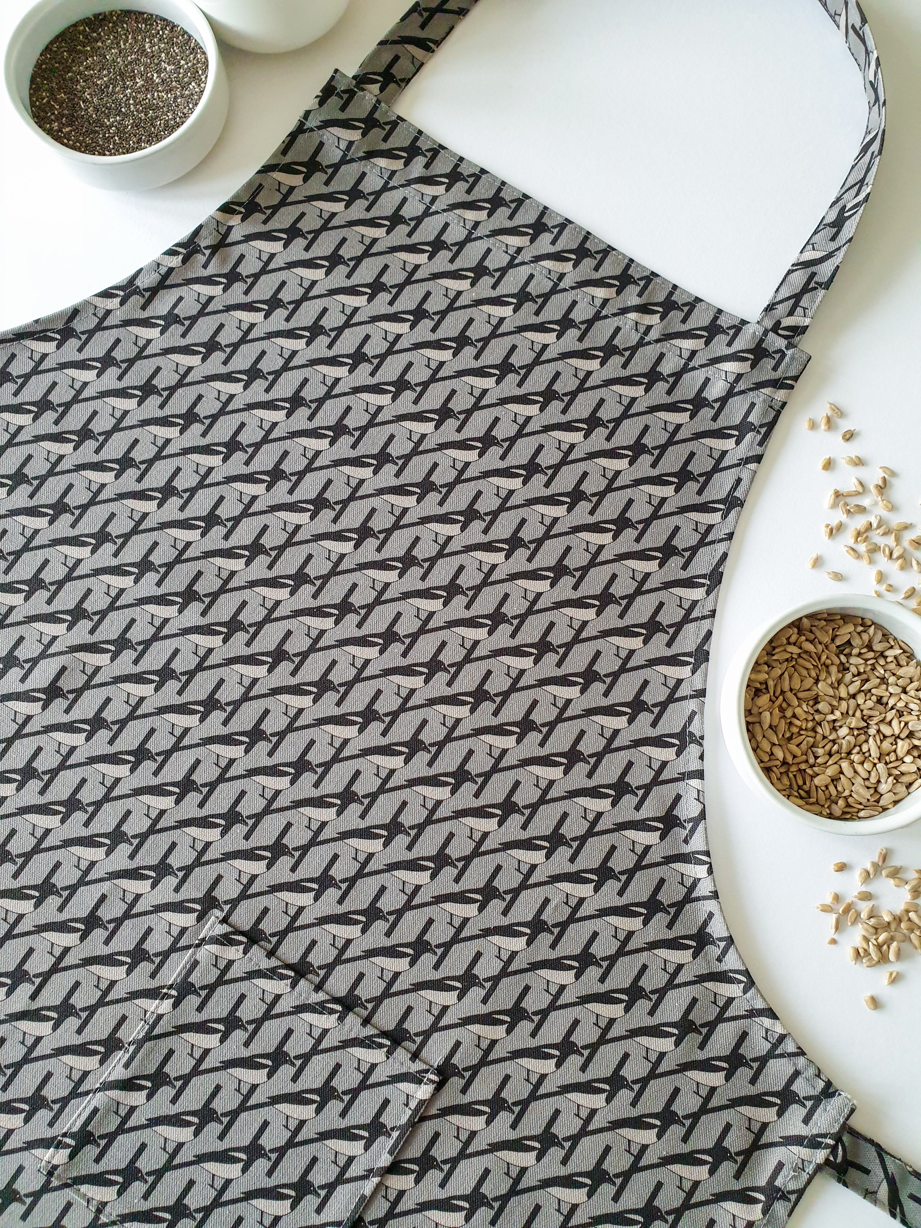 Organic cotton grey magpie apron – designed by Hannah Issi and handmade in the UK. Stylish, sustainable kitchen accessory - pictured on white background with cooking ingredients.
