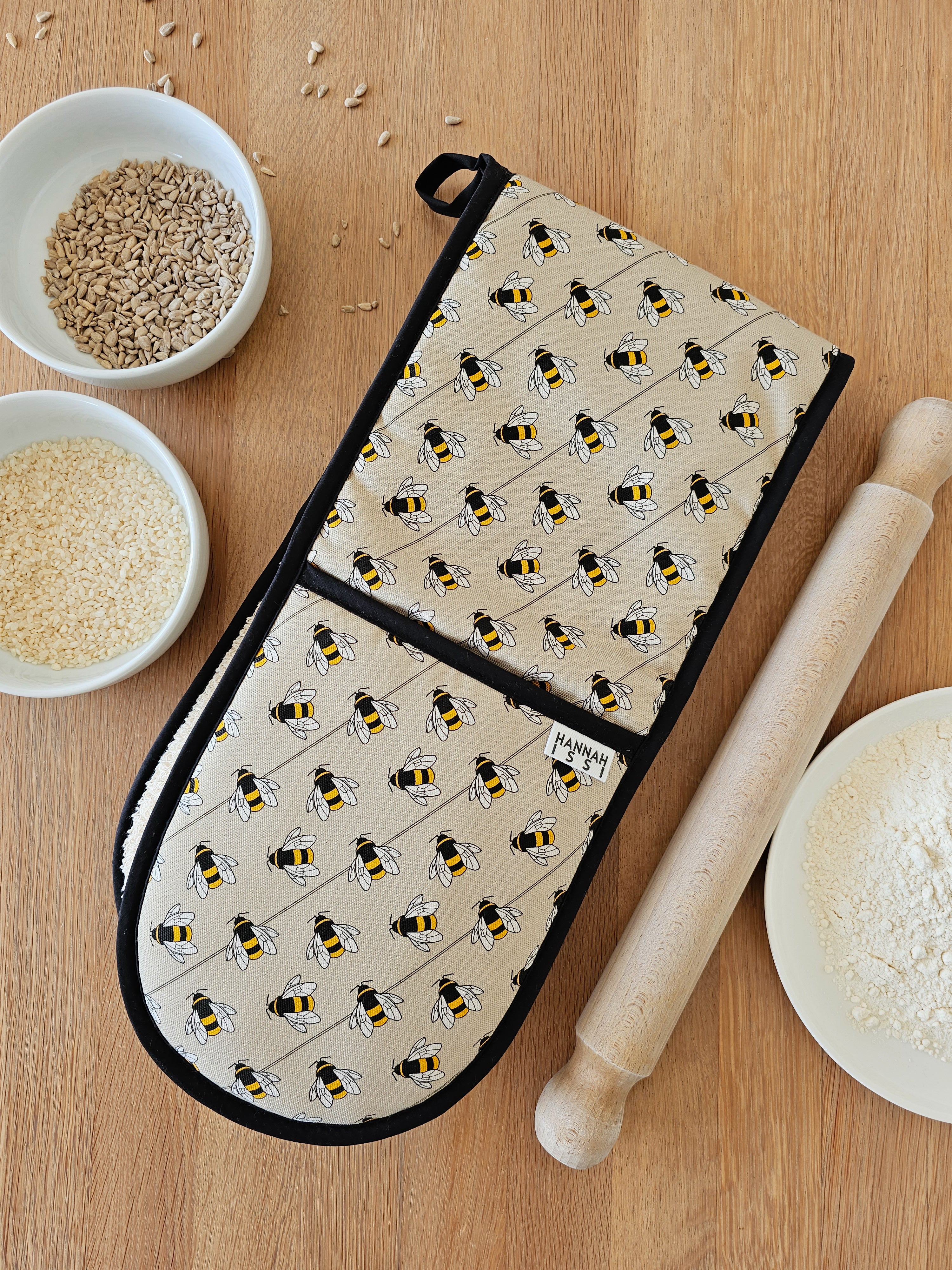 Organic cotton bee double oven glove – handmade in the UK by Hannah Issi. Stylish, heat-safe kitchen accessory. Image shows the glove on wooden tabletop with cooking ingredients.