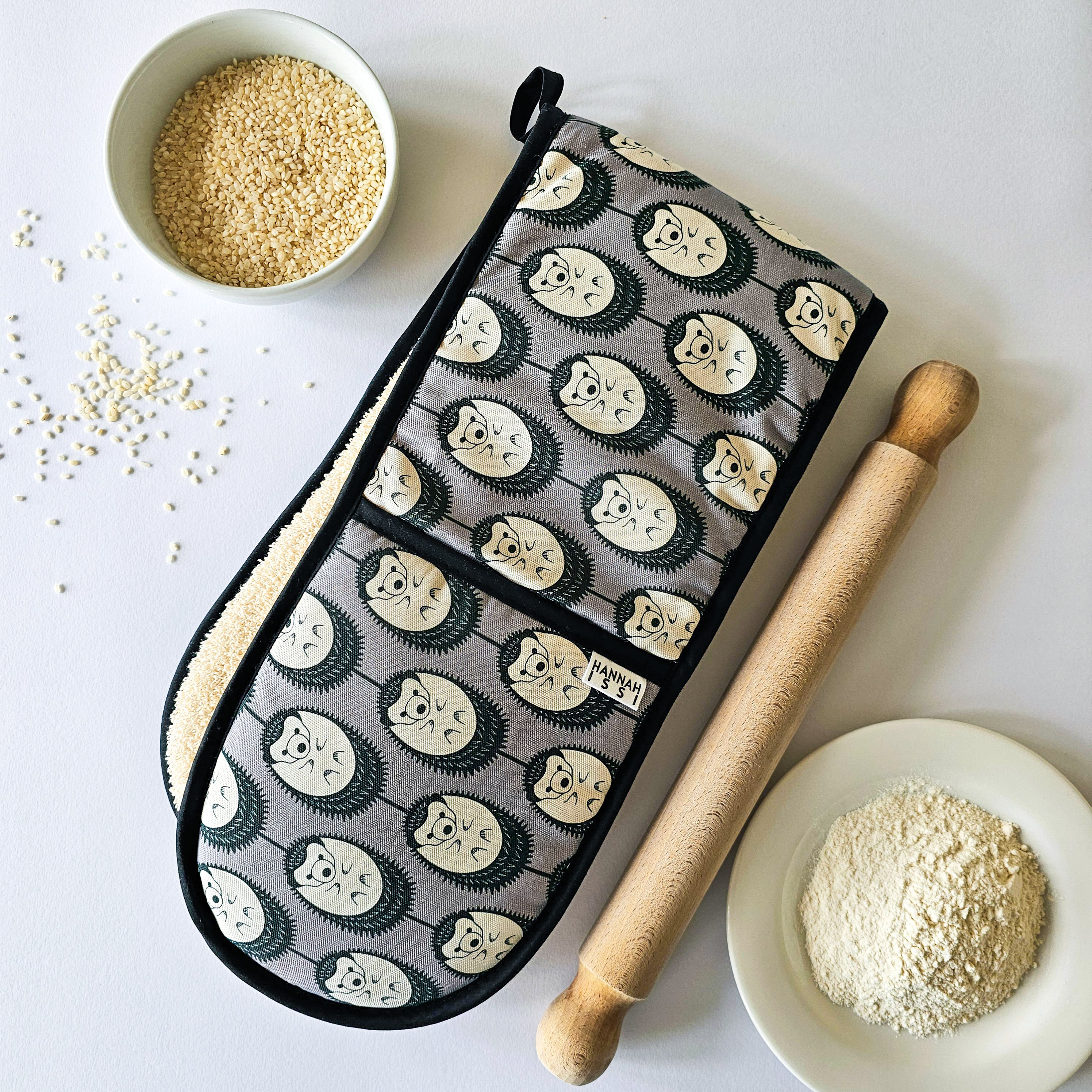 Organic cotton grey & white hedgehog double oven glove – handmade in the UK by Hannah Issi. Stylish, heat-safe kitchen accessory. Lifestyle image with cooking ingredients and rolling pin on white background.
