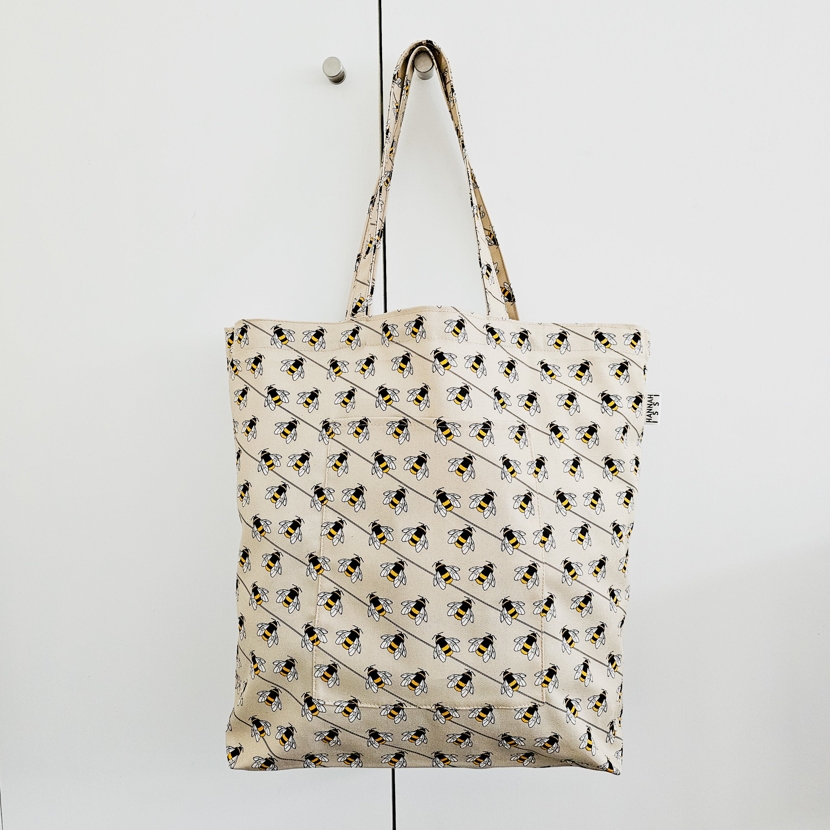 Organic cotton bee shoulder bag – handmade in the UK by Hannah Issi. Large, sustainable accessory for every day. Bag shown hanging from doorknob on white cupboard.