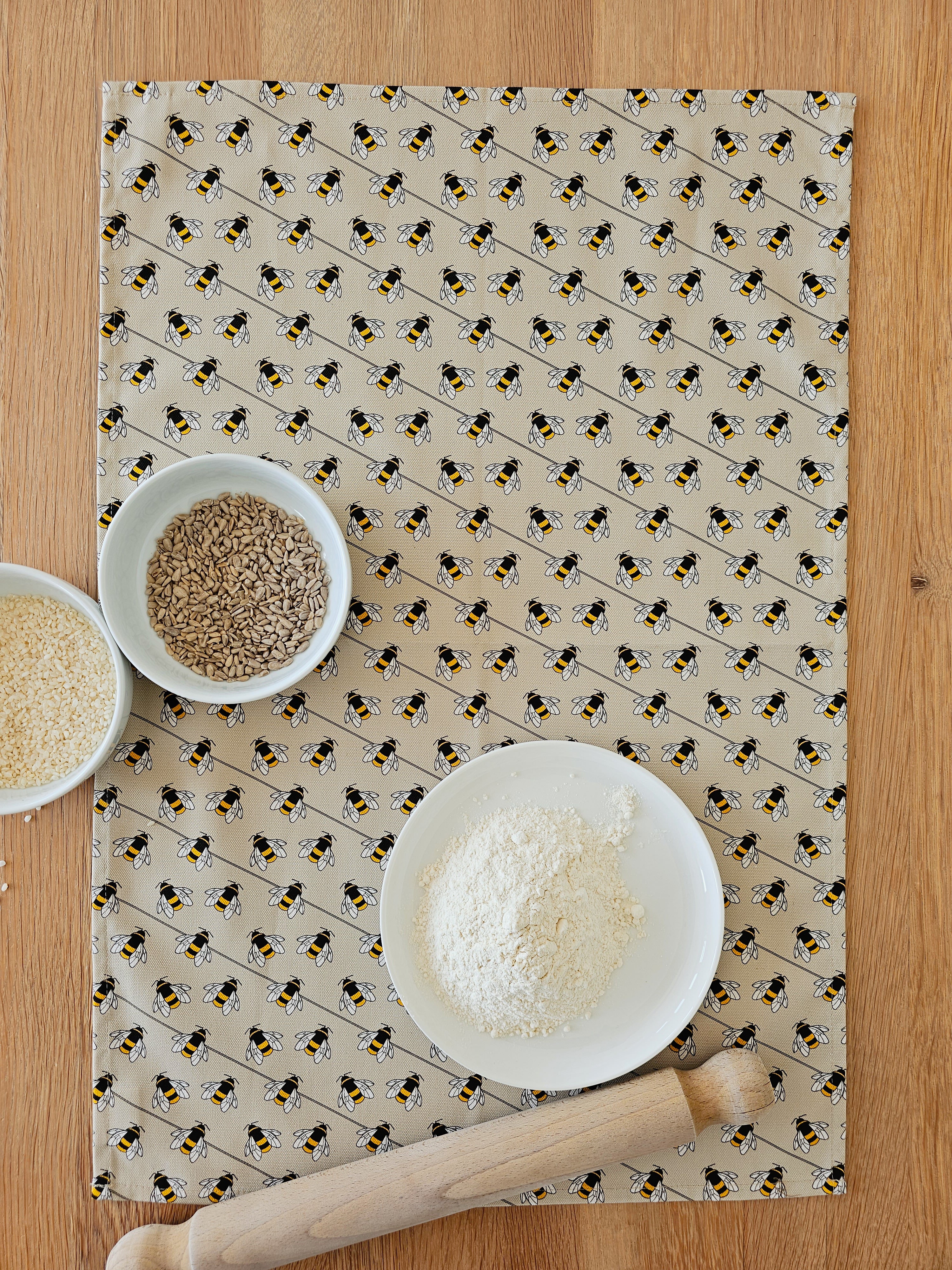 Organic cotton bee tea towel – designed by Hannah Issi and handmade in the UK. Stylish, sustainable kitchen linen. Shown laying on wooden table with cooking ingredients.