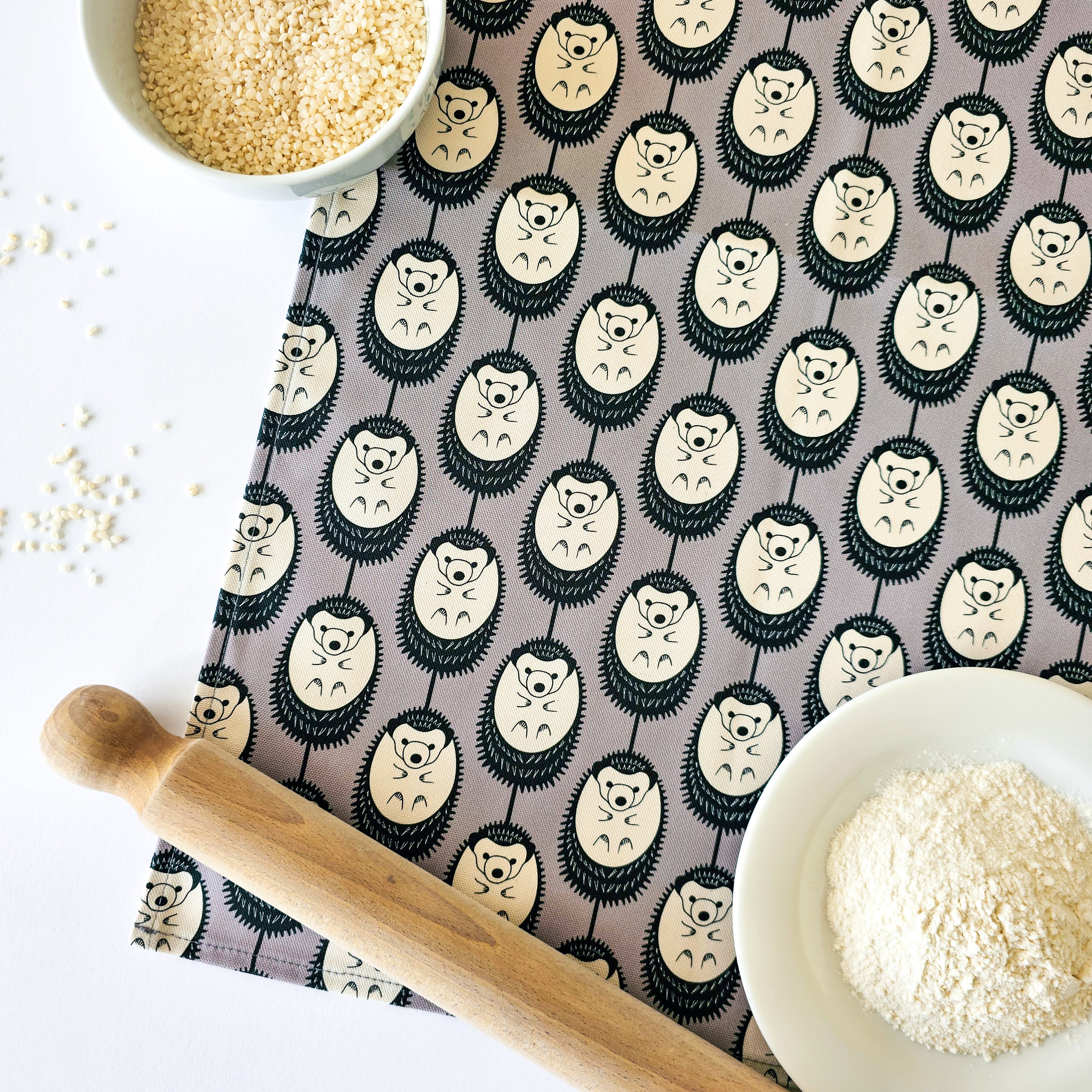 Organic cotton grey and white hedgehog tea towel – designed by Hannah Issi and handmade in the UK. Stylish, sustainable kitchen linen- shown on white background with wooden rolling pin and cooking ingredients.