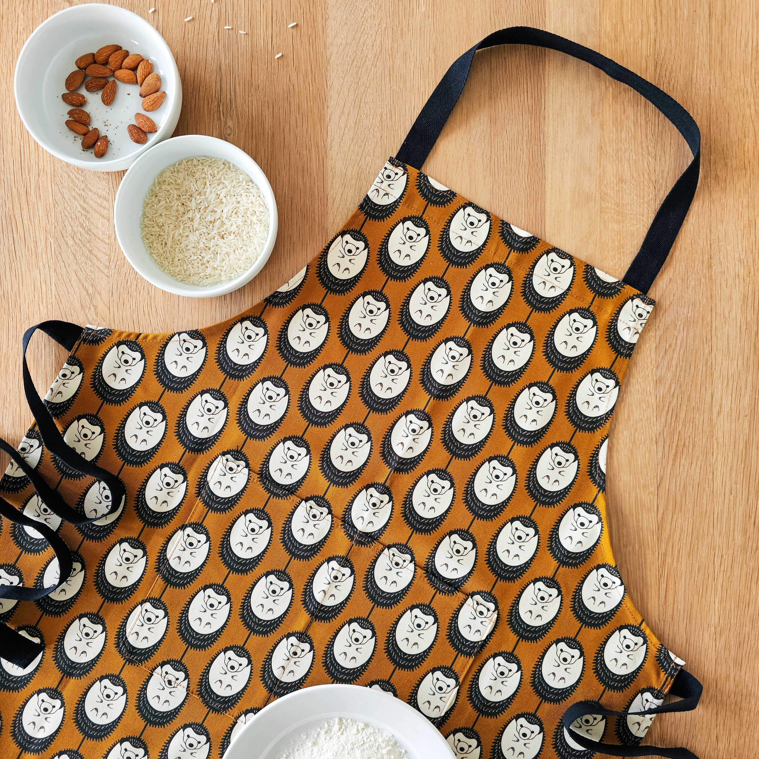 Organic cotton brown and white hedgehog apron – designed by Hannah Issi and handmade in the UK. Stylish, sustainable kitchen textile inspired by nature. Lifestyle photo shows apron lying on wooden table with cooking ingredients. 