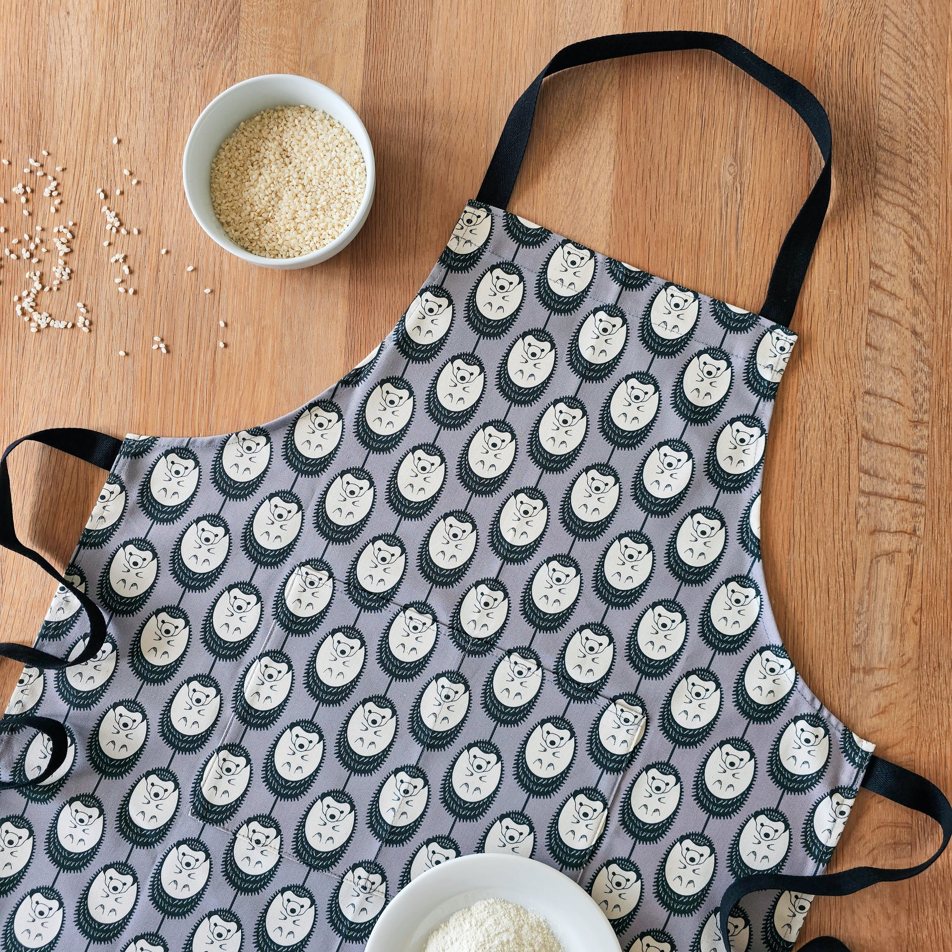 Organic cotton grey and white hedgehog apron – designed by Hannah Issi and handmade in the UK. Stylish, sustainable kitchen textile. Lifestyle photo showing apron laying on wooden table with cooking ingredients. 