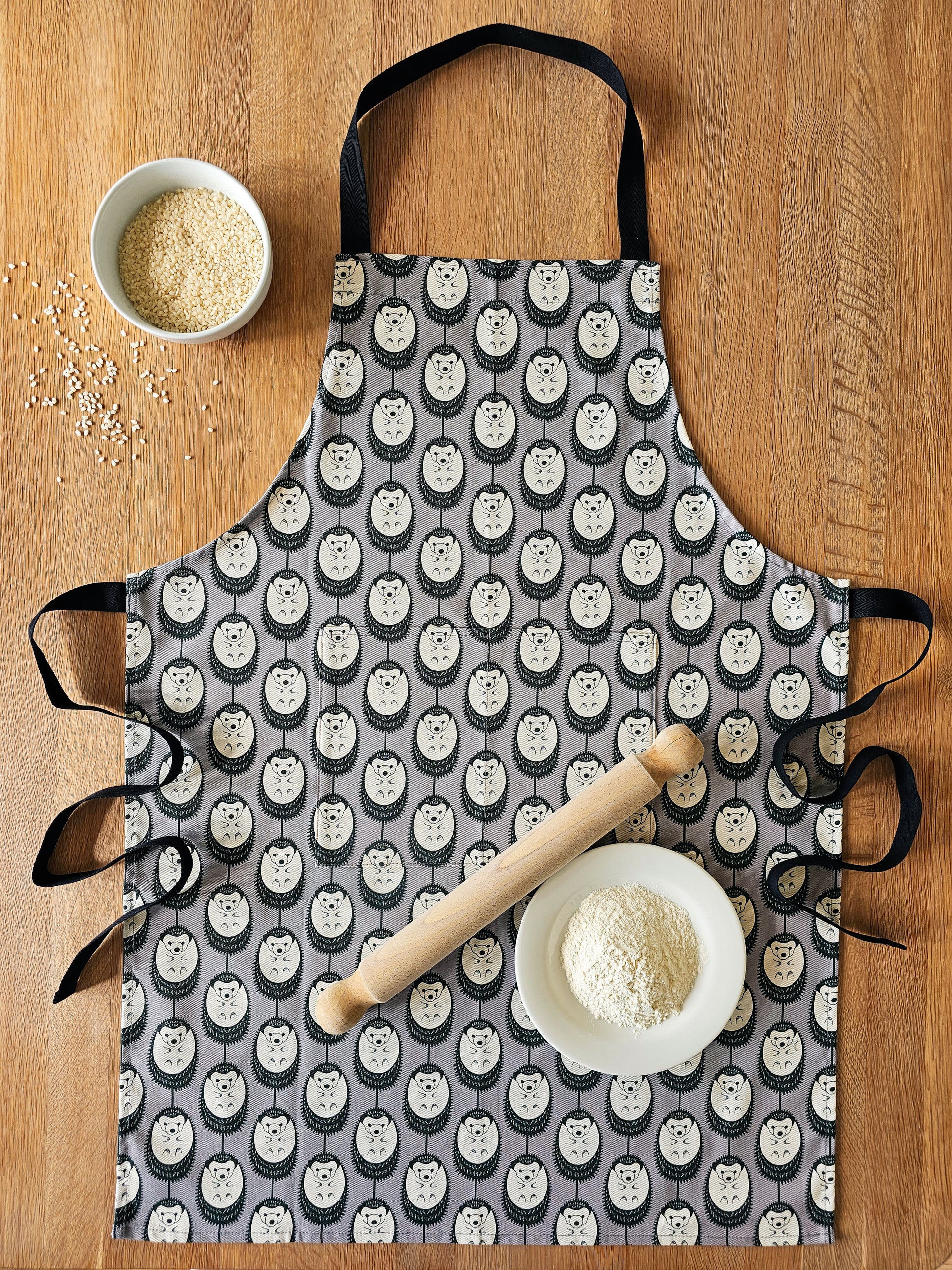 Organic cotton grey and white hedgehog apron – designed by Hannah Issi and handmade in the UK. Stylish, sustainable kitchen textile. Photo shows apron laying on wooden table with cooking ingredients. 