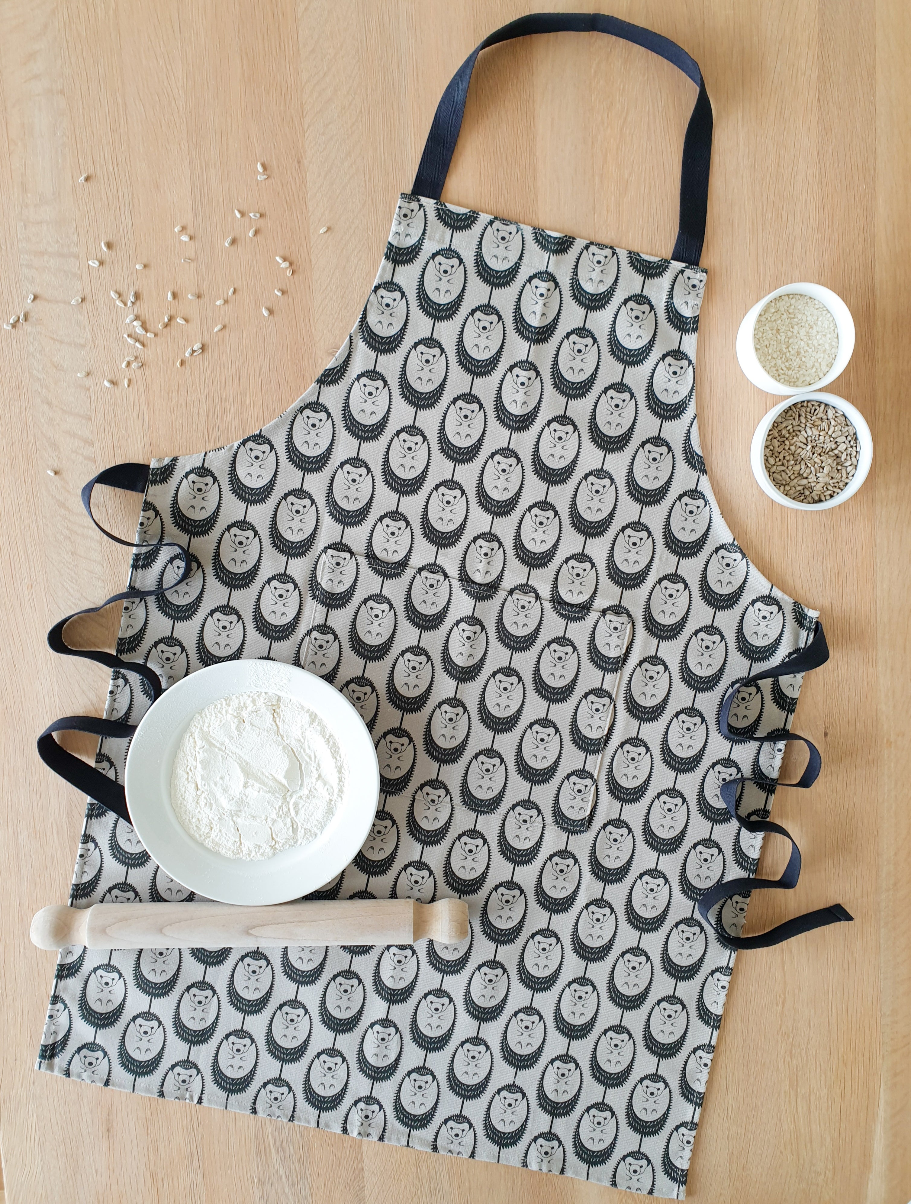 Organic cotton grey hedgehog apron – designed by Hannah Issi and handmade in the UK. Stylish, sustainable kitchen textile. Pictured laying on wooden table with cooking ingredients.
