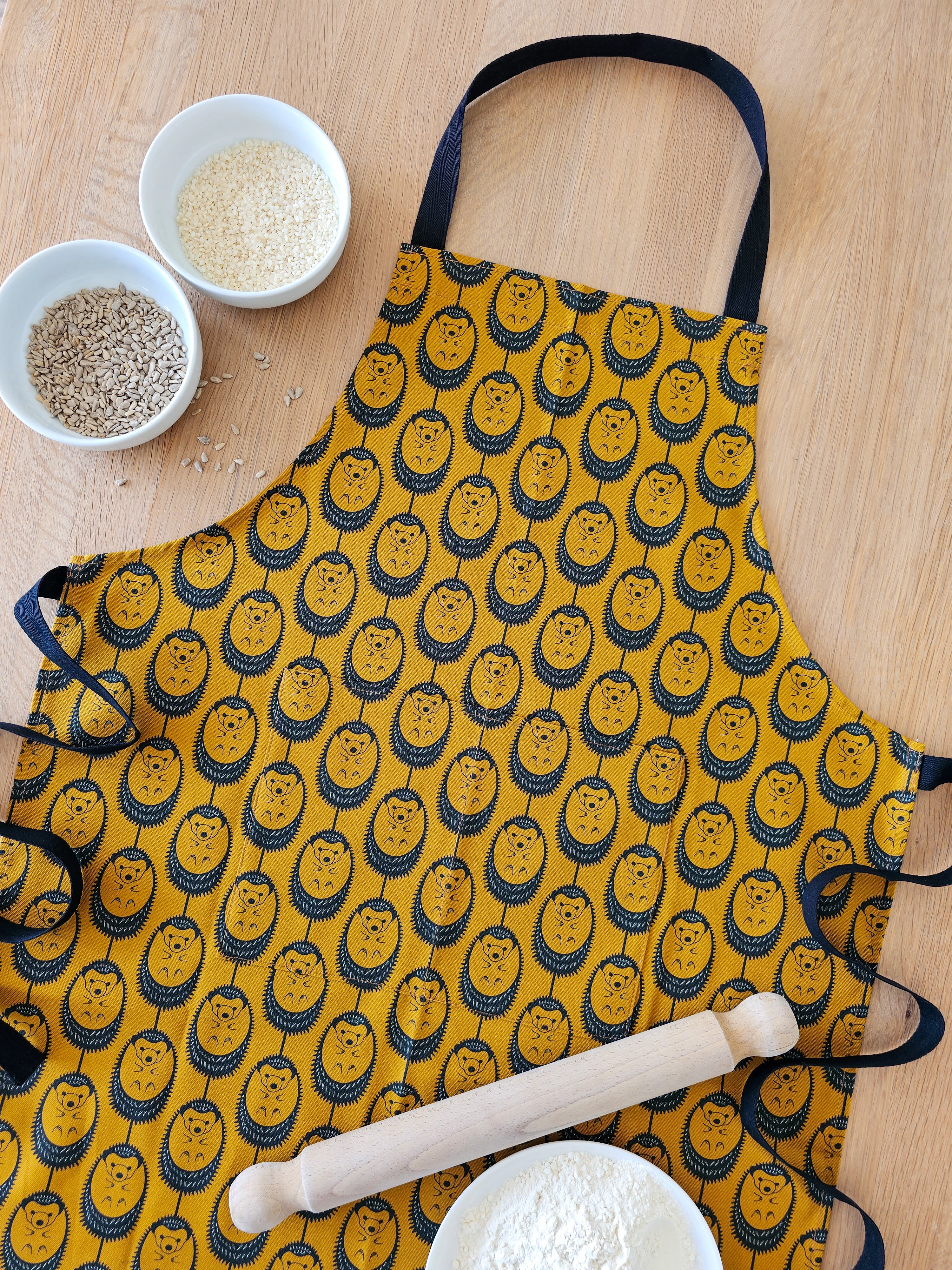 Organic cotton ochre hedgehog apron – designed by Hannah Issi and handmade in the UK. Stylish, sustainable kitchen textile, pictured laying on wooden table with cooking ingredients.