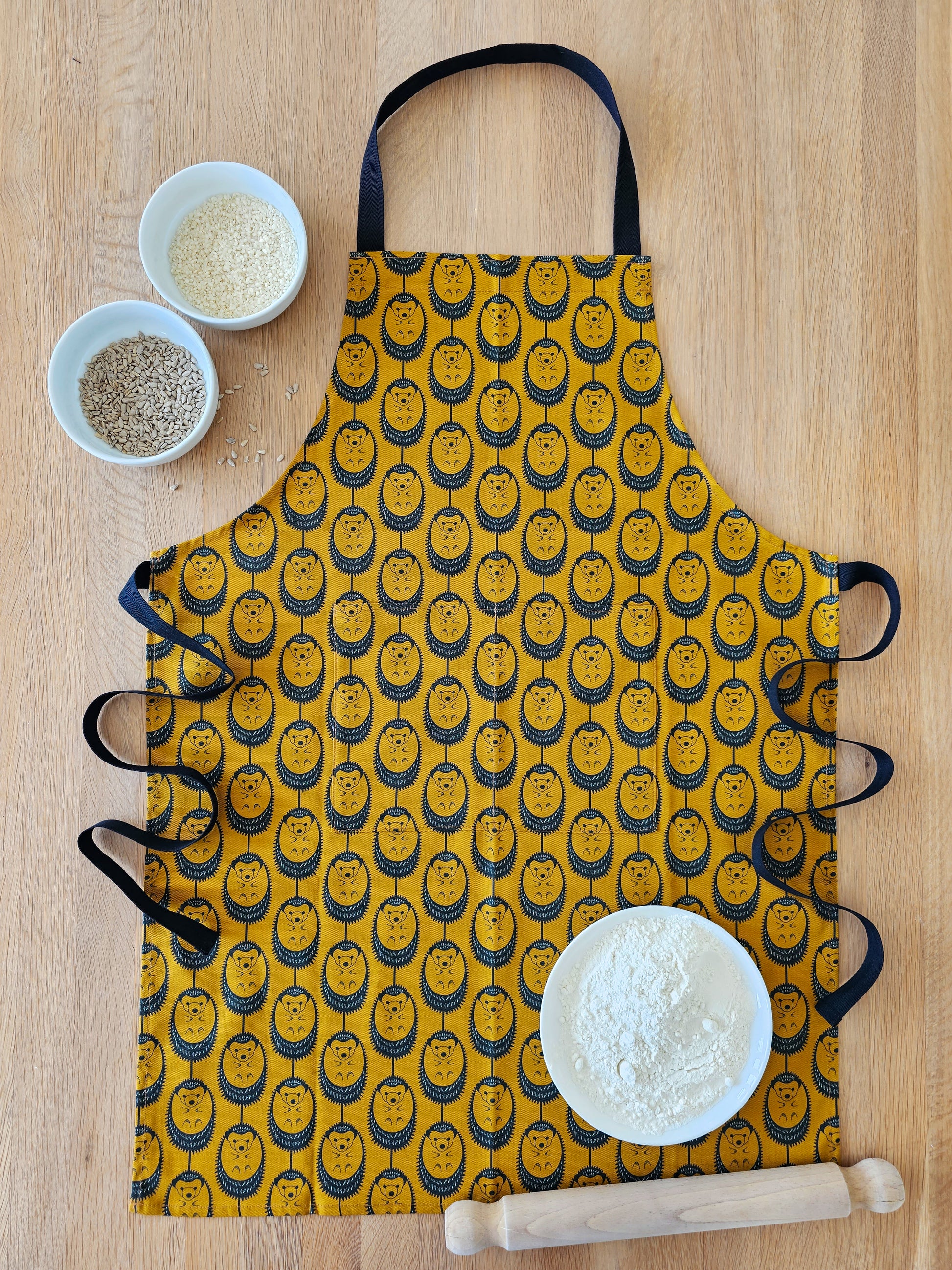 Organic cotton ochre hedgehog apron – designed by Hannah Issi and handmade in the UK. Stylish, sustainable kitchen textile- lifestyle shot pictured with wooden background and cooking ingredients. 