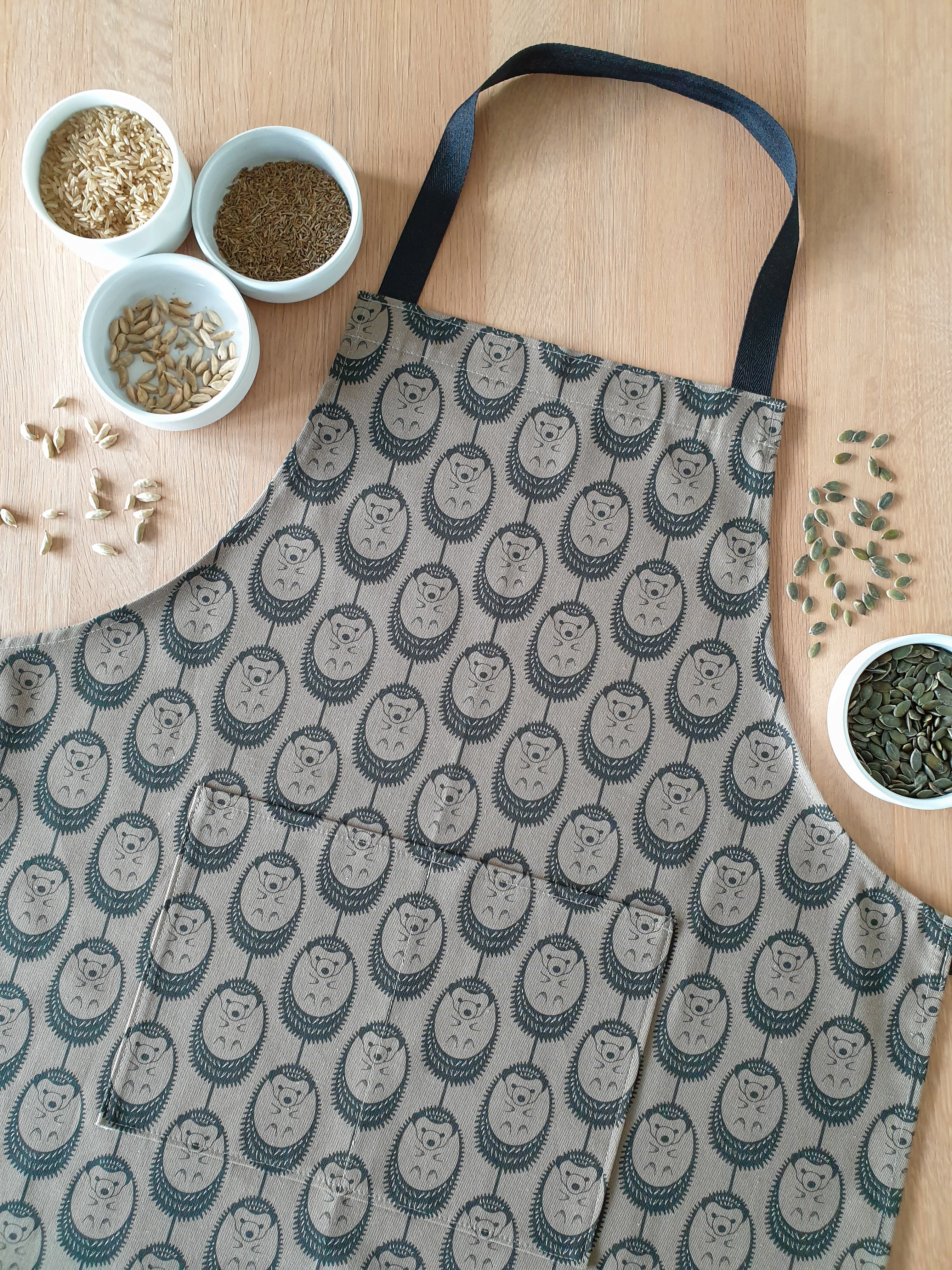 Organic cotton olive green hedgehog apron – designed by Hannah Issi and handmade in the UK. Stylish, sustainable kitchen accessory pictured with cooking ingredients.