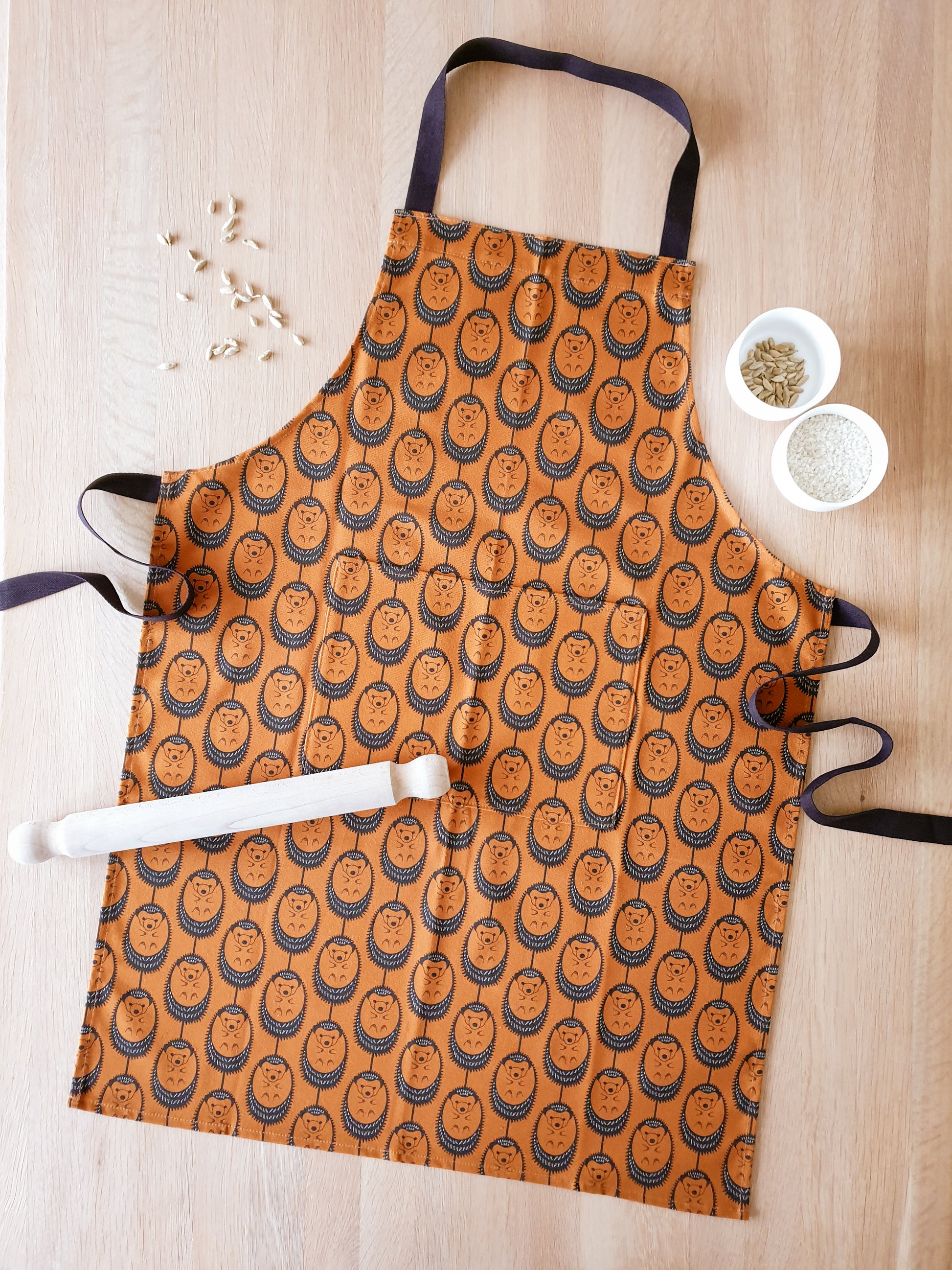 Organic cotton terracotta hedgehog apron – designed by Hannah Issi and handmade in the UK. Sustainable, modern kitchen textile pictured on wooden table with cooking ingredients.