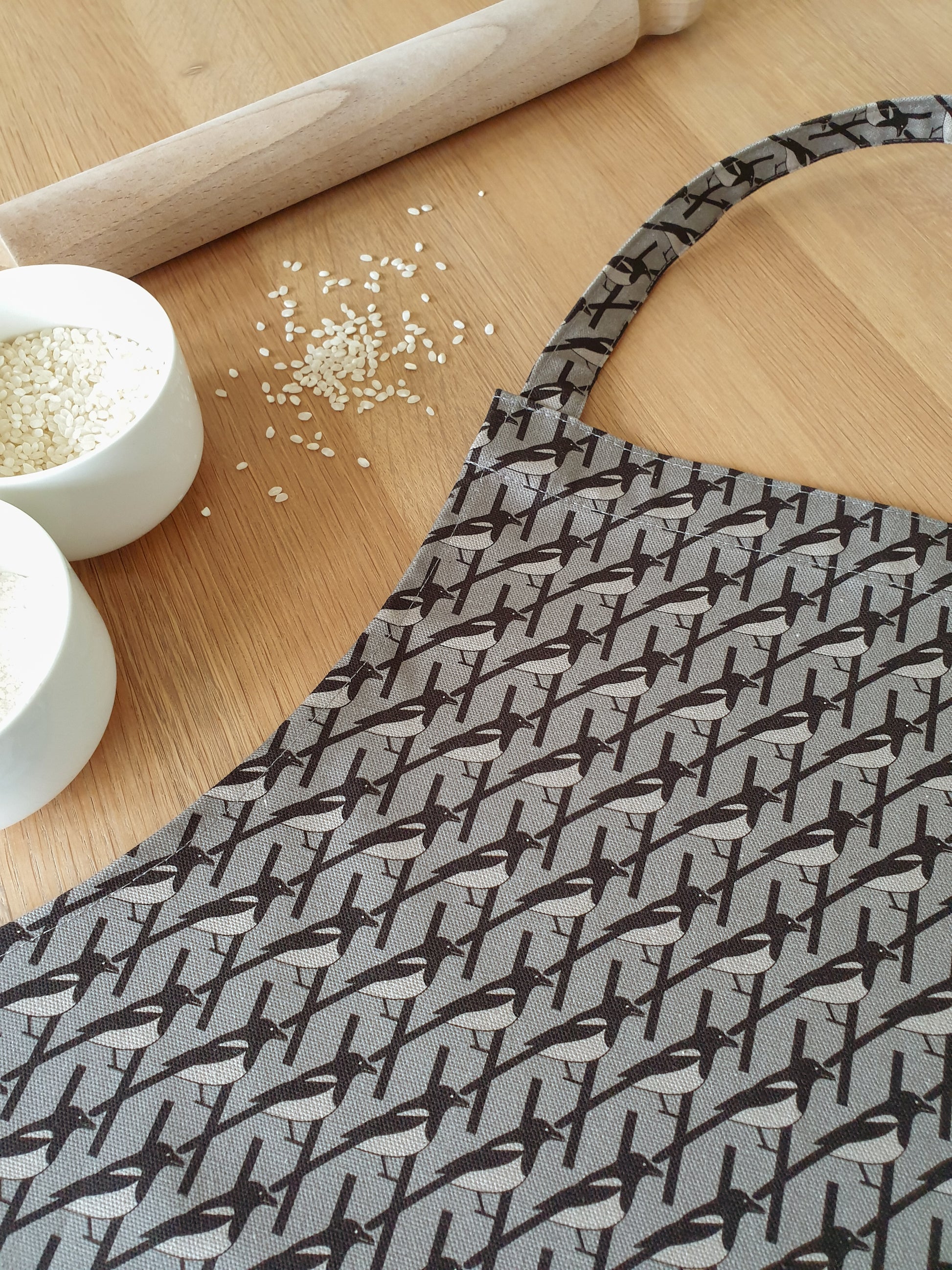 Organic cotton grey magpie apron – designed by Hannah Issi and handmade in the UK. Stylish, sustainable kitchen accessory - photo shows close-up of print with wooden background and cooking ingredients.