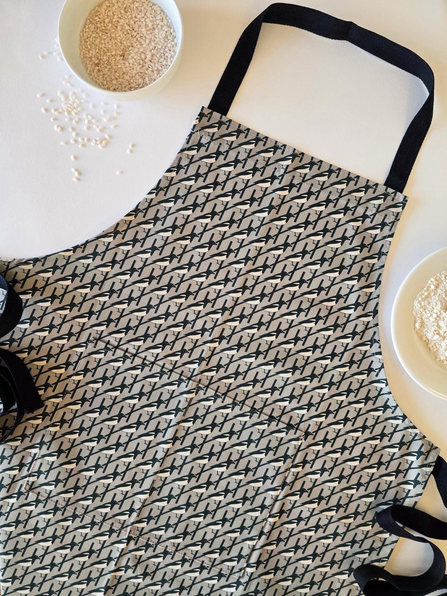 Organic cotton grey magpie apron – designed by Hannah Issi and handmade in the UK. Stylish, sustainable kitchen accessory - close-up picture on white background with cooking ingredients.