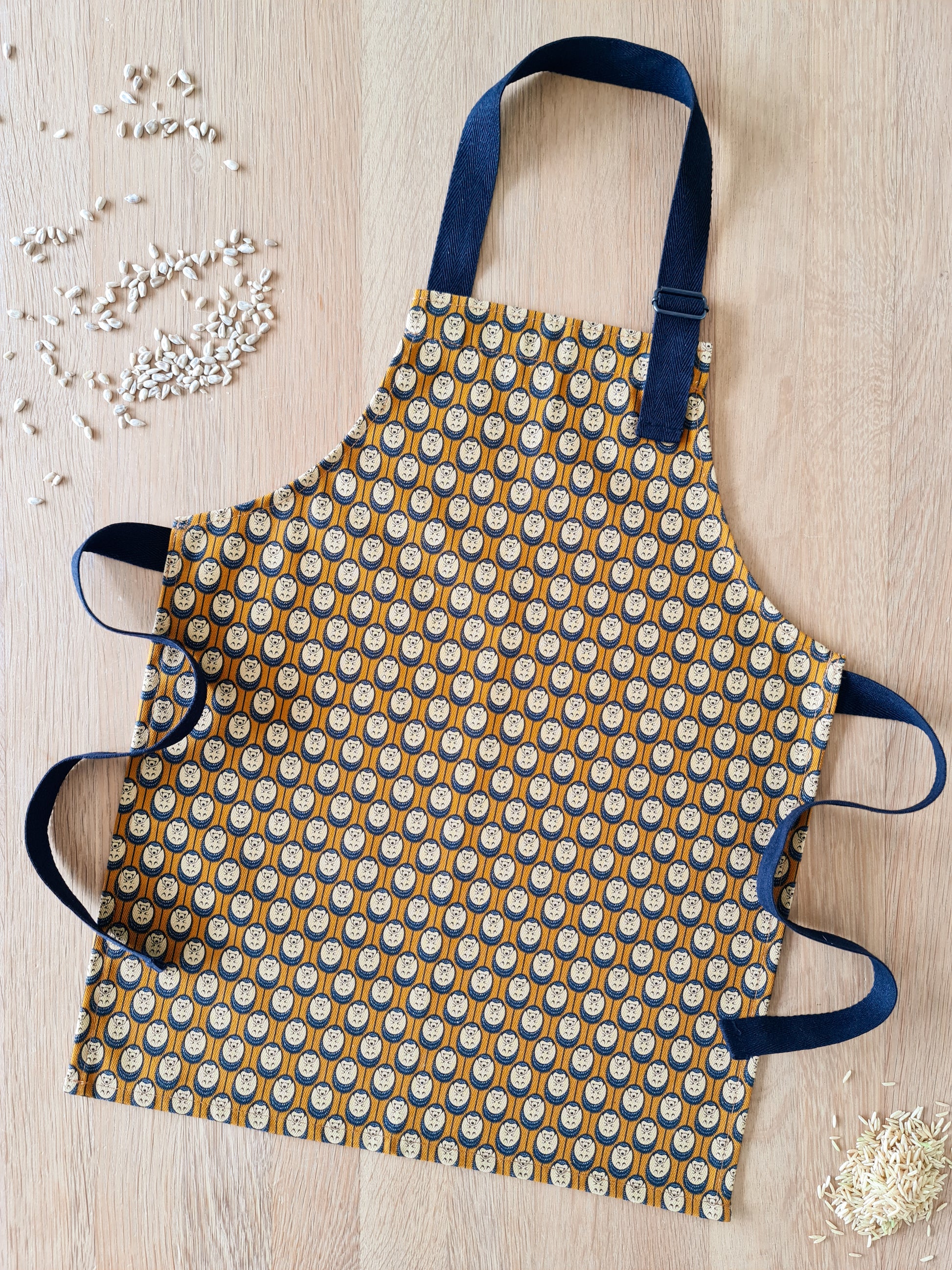 Organic cotton autumn hedgehog apron set (adult & child) – handmade in the UK by Hannah Issi. Stylish, sustainable kitchen pair- shows children's apron on wooden tabletop with cooking ingredients.