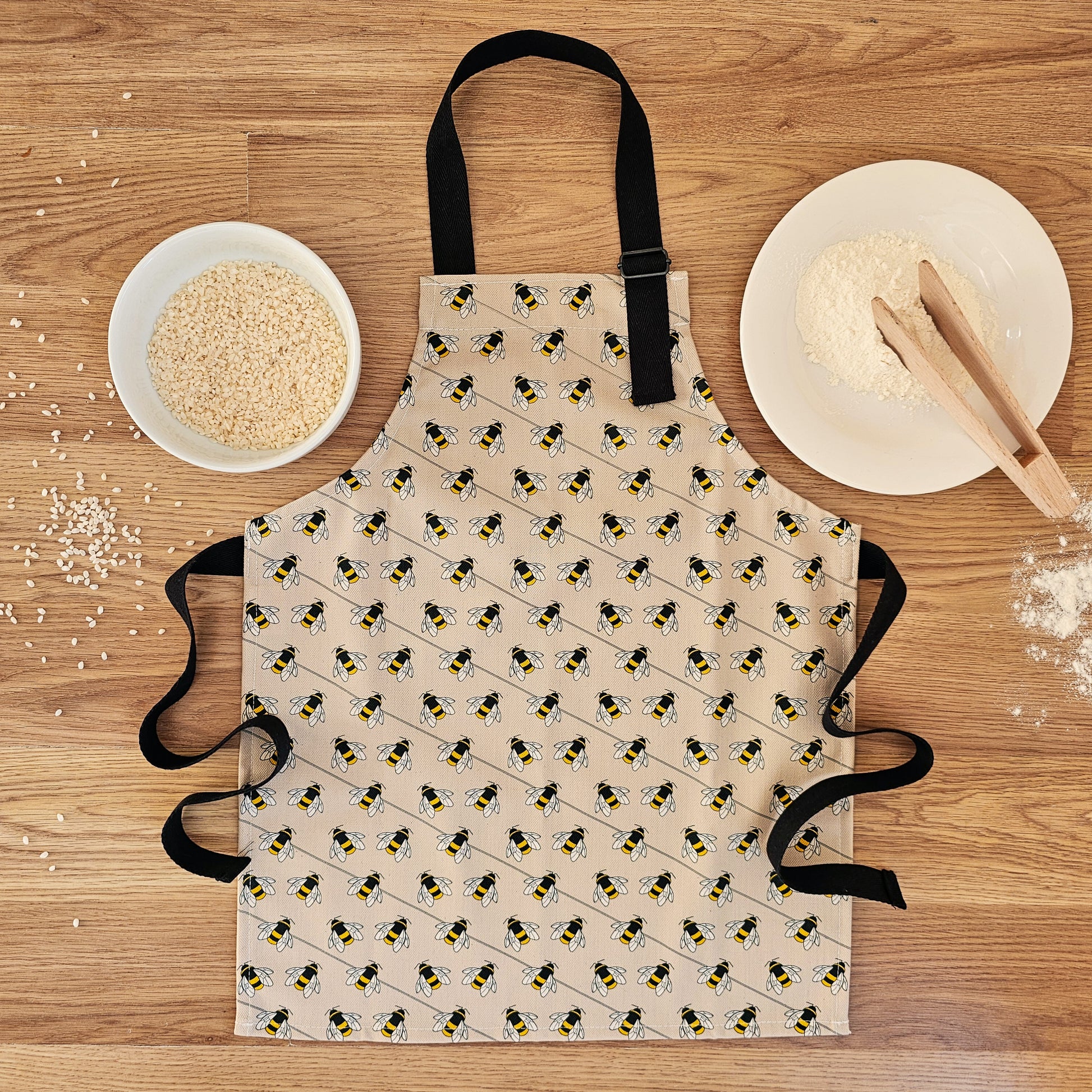 Organic cotton children’s bee apron – handmade in the UK by Hannah Issi. Fun, sustainable kitchen accessory for kids for cooking or messy play. Image shows apron laying on wooden table with cooking ingredients. 
