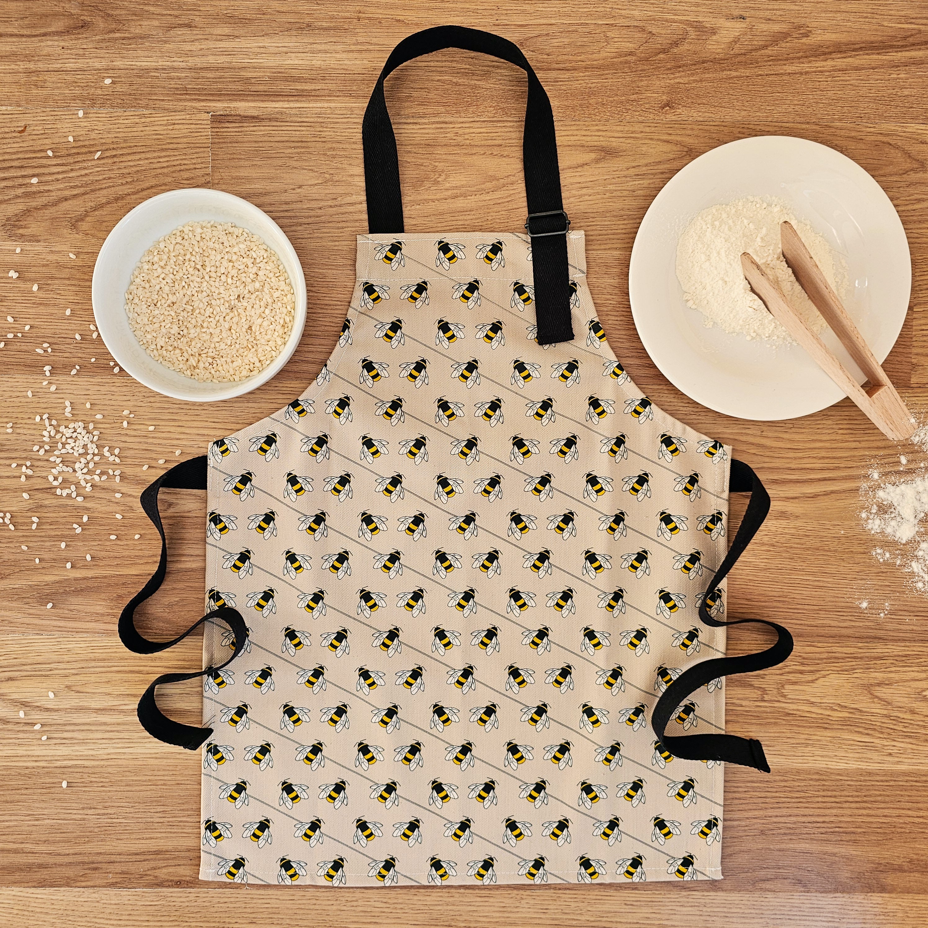 Organic cotton children’s bee apron – handmade in the UK by Hannah Issi. Fun, sustainable kitchen accessory for kids for cooking or messy play. Image shows apron laying on wooden table with cooking ingredients. 