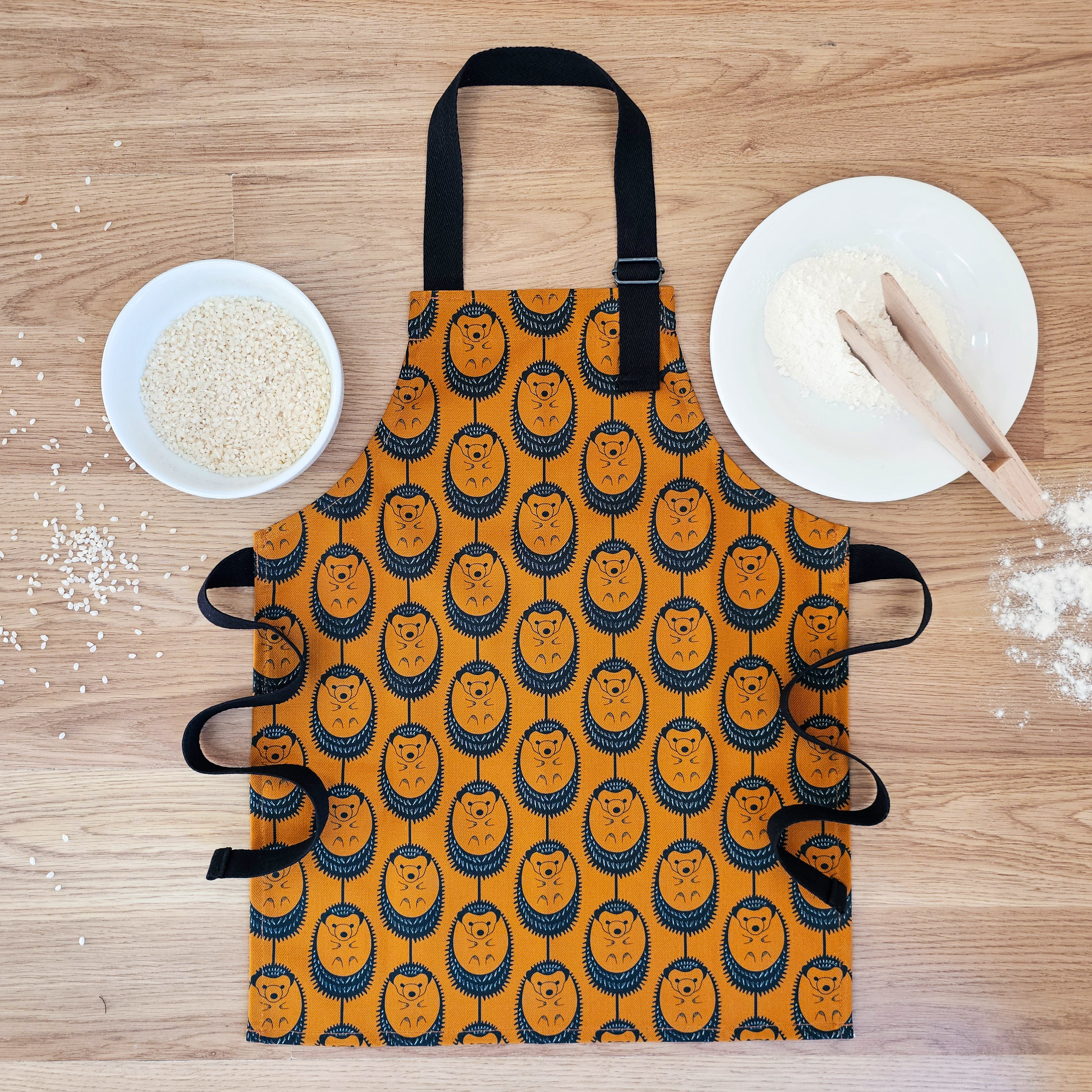 Organic cotton children’s terracotta hedgehog apron – handmade in the UK by Hannah Issi. Fun, sustainable kitchen accessory for kids. Lifestyle image - apron laying on wooden table with cooking ingredients.