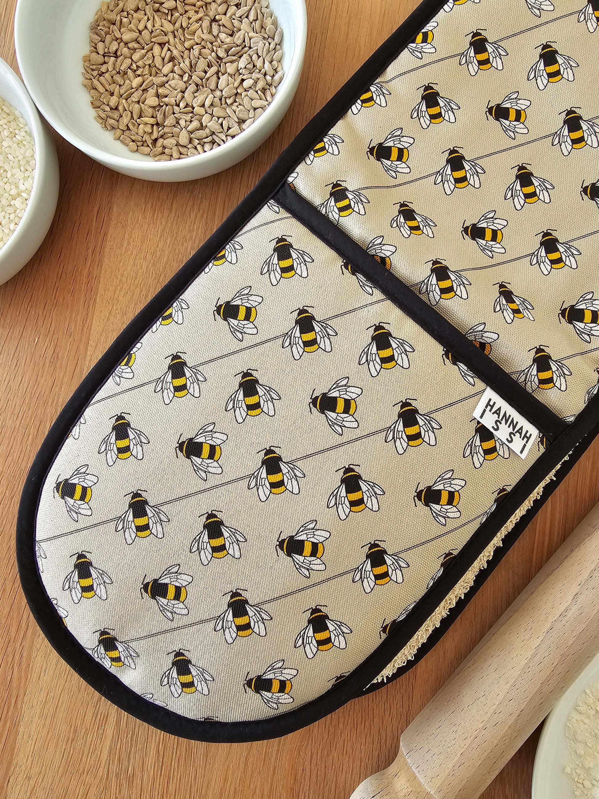 Organic cotton bee double oven glove – handmade in the UK by Hannah Issi. Stylish, heat-safe kitchen accessory. Image shows close-up  on wooden tabletop with cooking ingredients. 