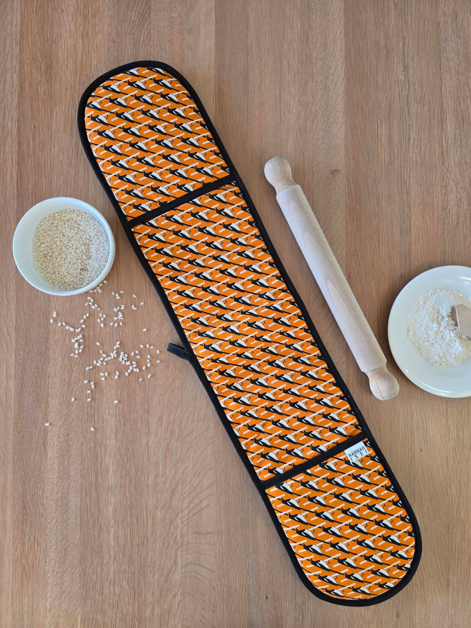 Organic cotton orange magpie double oven glove – handmade in the UK by Hannah Issi. Stylish, heat-safe kitchen accessory. Lifestyle image showing oven glove laid out on wooden tabletop with cooking ingredients.
