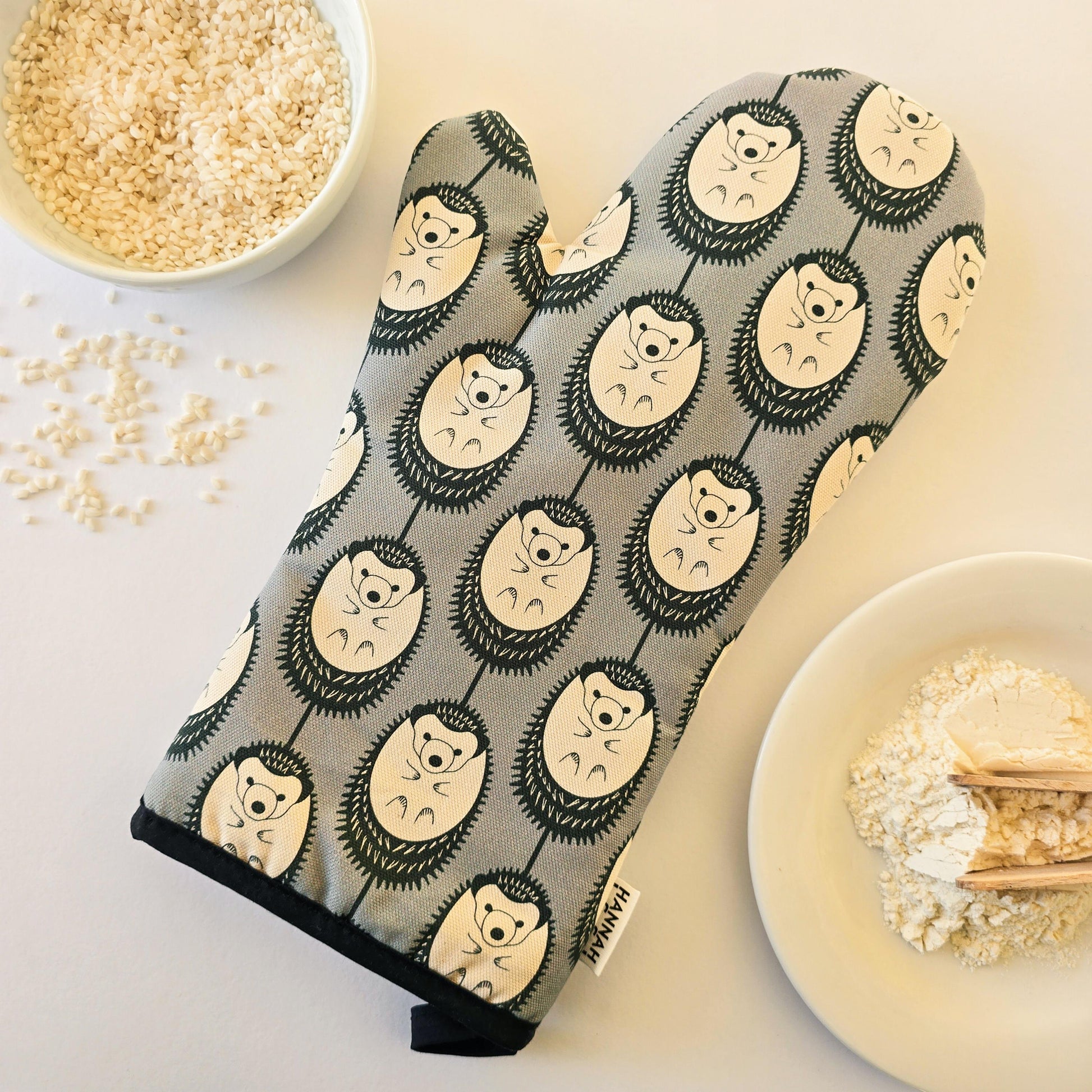 Organic cotton grey & white hedgehog luxury oven glove – handmade in the UK by Hannah Issi. Stylish, heat-safe kitchen accessory. Lifestyle image with white background and cooking ingredients.