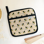 Organic cotton Bee pot grab – heat-safe kitchen handle cover by Hannah Issi, handmade in the UK. Stylish and functional. Image shown with cooking accessories on white background.