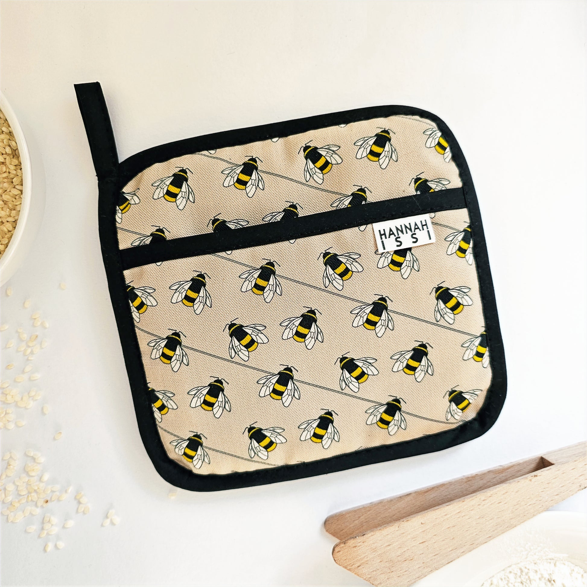Organic cotton Bee pot grab – heat-safe kitchen handle cover by Hannah Issi, handmade in the UK. Stylish and functional. Image shown with cooking accessories on white background.