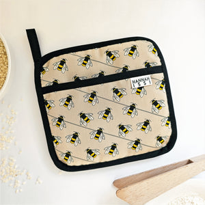 Organic cotton Bee pot grab – heat-safe kitchen handle cover by Hannah Issi, handmade in the UK. Stylish and functional. Image shown with cooking accessories on white background.