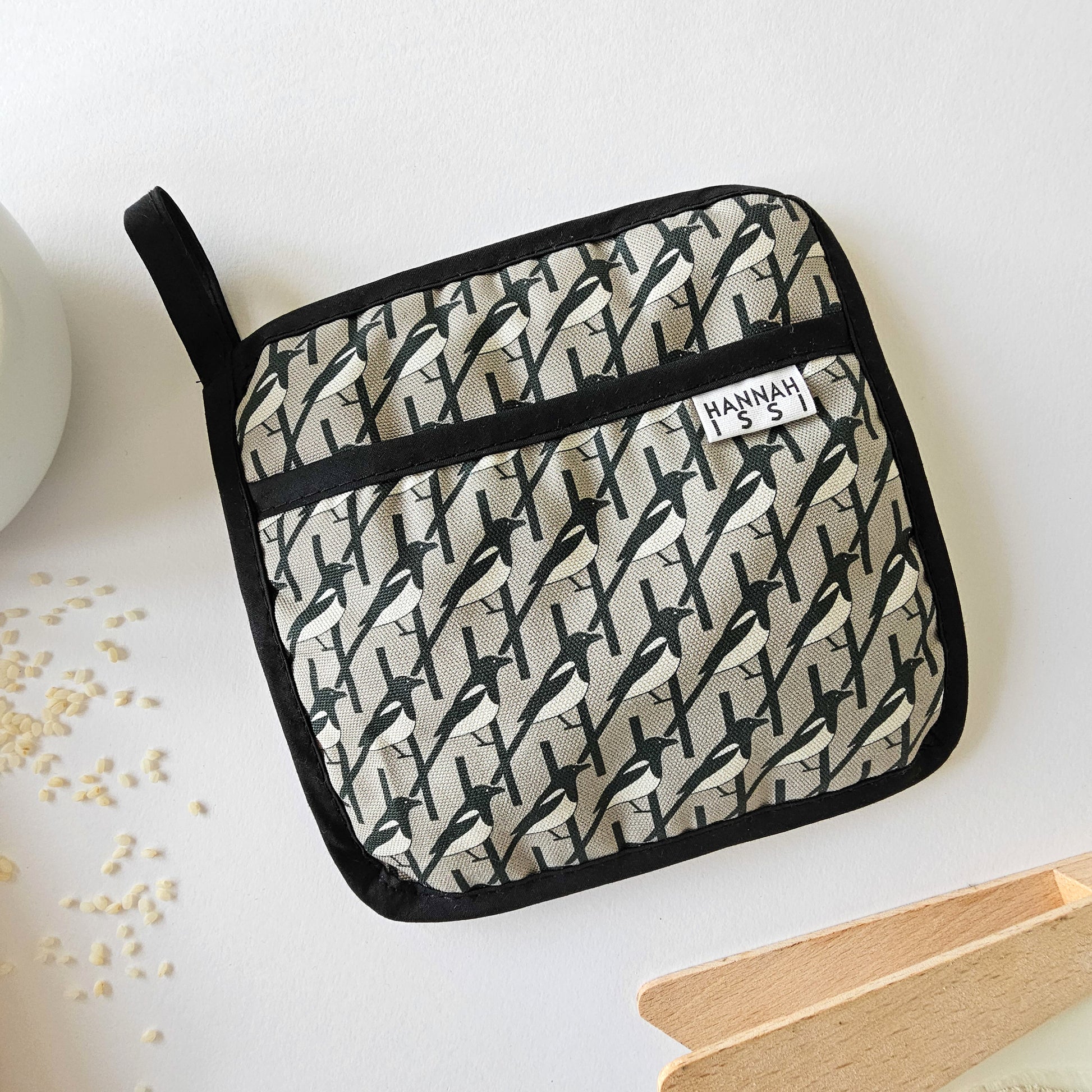 Organic cotton Grey Magpie pot grab – heat-safe kitchen handle cover by Hannah Issi, handmade in the UK. Stylish and functional. Image is on white background with kitchen accessories. 