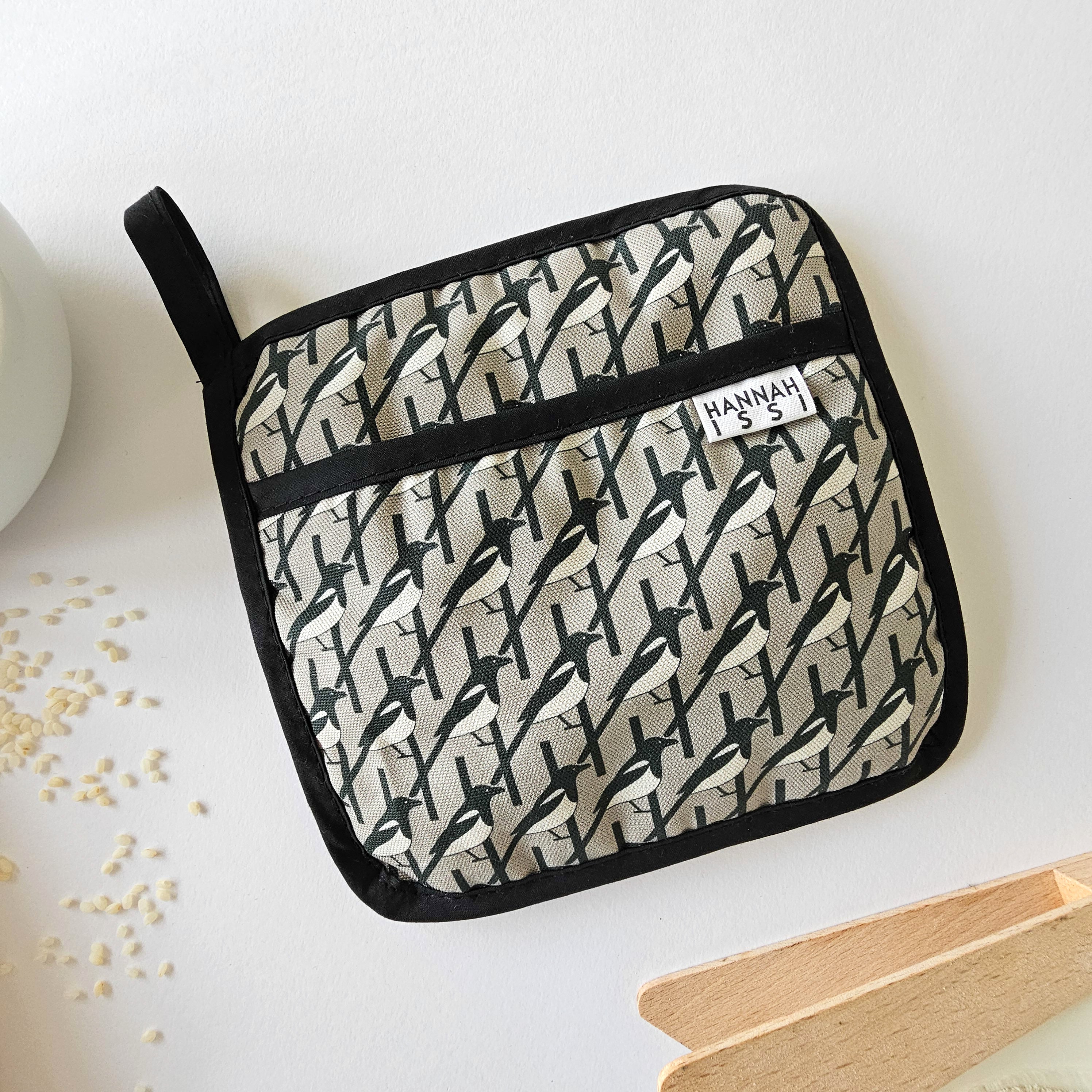 Organic cotton Grey Magpie pot grab – heat-safe kitchen handle cover by Hannah Issi, handmade in the UK. Stylish and functional. Image is on white background with kitchen accessories. 