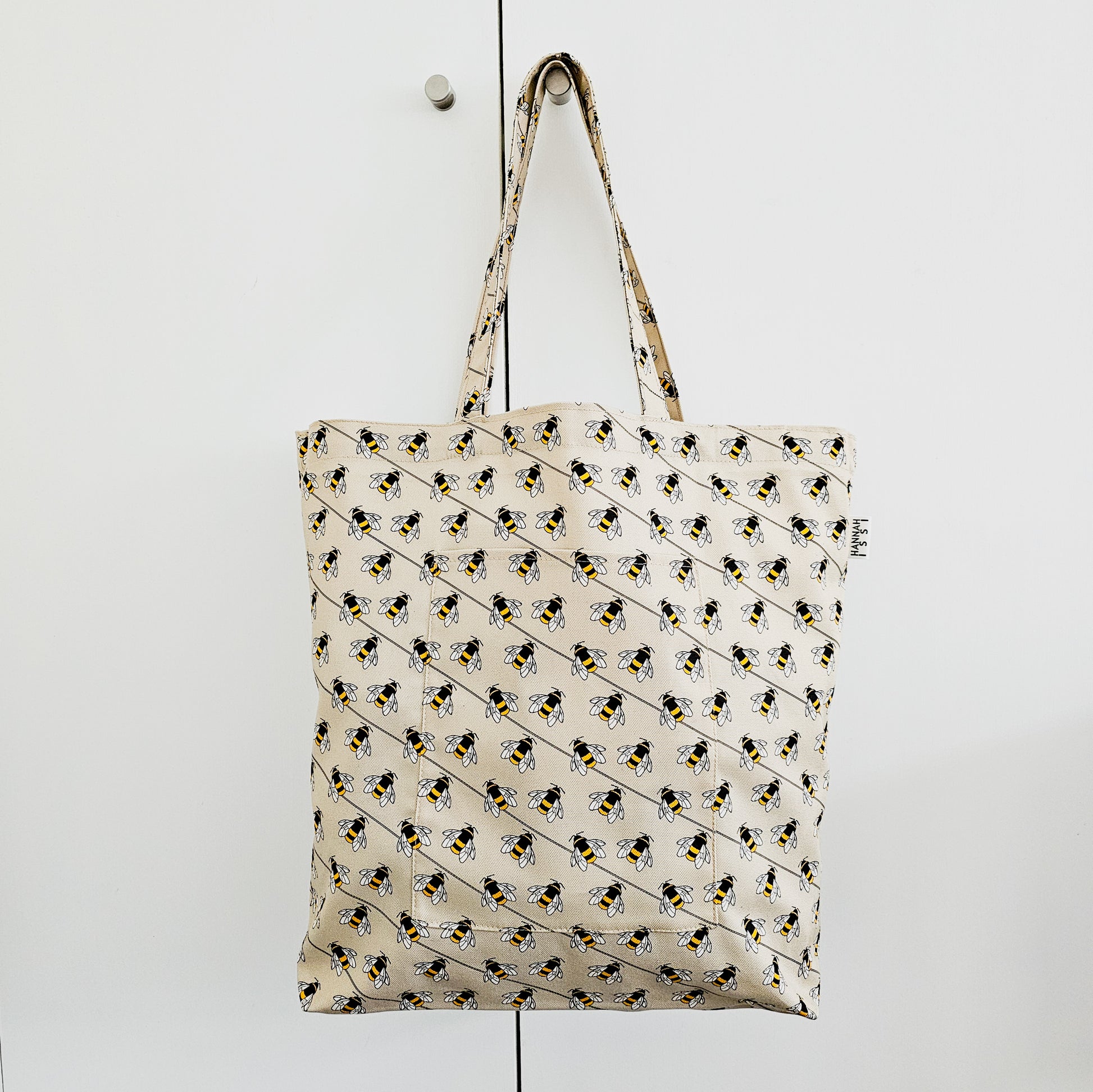 Organic cotton bee shoulder bag – handmade in the UK by Hannah Issi. Large, sustainable accessory for every day. Bag shown hanging from doorknob on white cupboard.