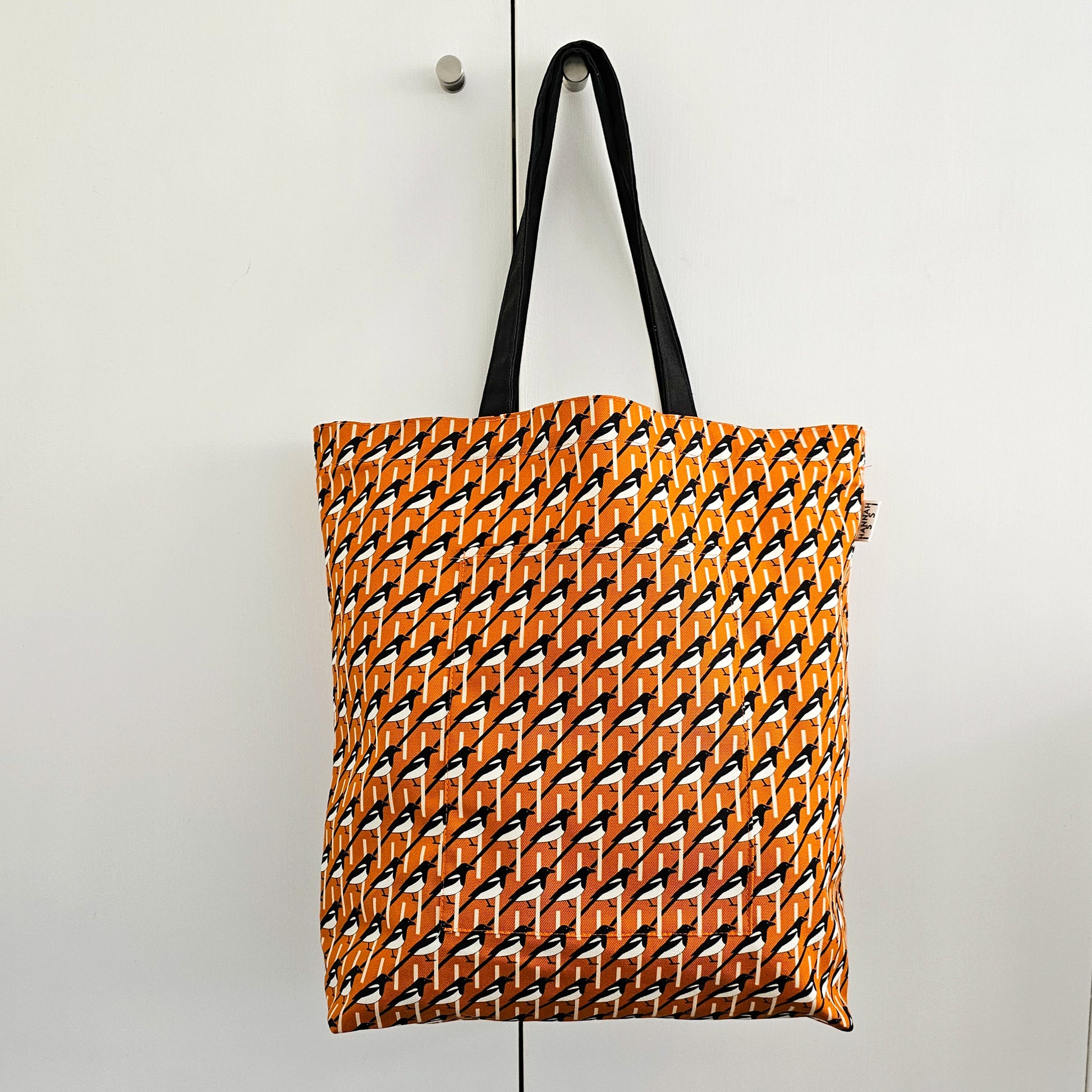 Organic cotton orange Magpie shoulder bag – handmade in the UK by Hannah Issi. Large, sustainable accessory for every day. Bag shown hanging from doorknob on white cupboard doors. 