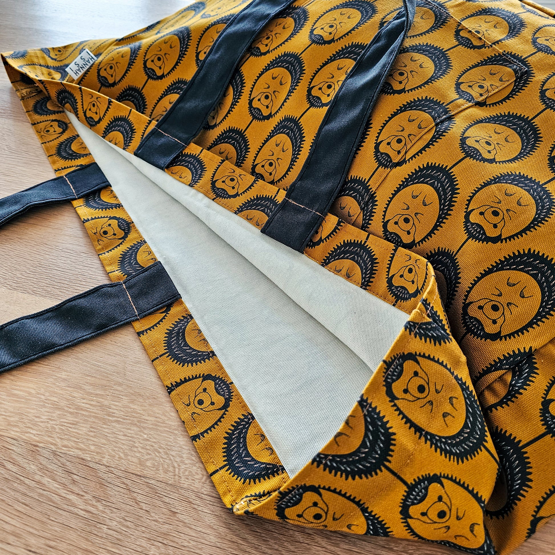 Organic cotton Ochre Hedgehog oversized shoulder bag – large, roomy design handmade in the UK by Hannah Issi. Stylish and sustainable. Shows close-up detail of bag print and shoulder straps on wooden tabletop.