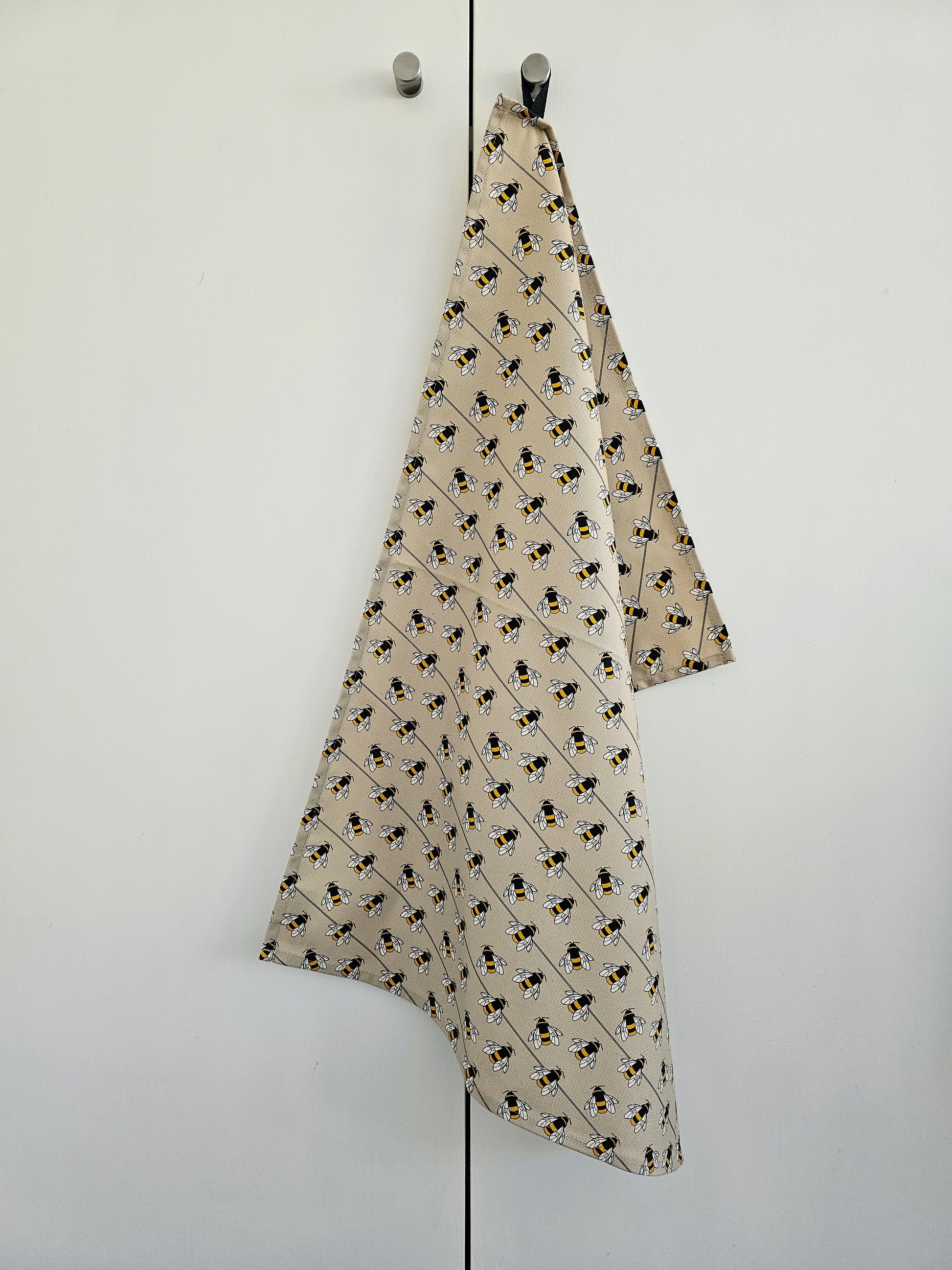 Organic cotton bee tea towel – designed by Hannah Issi and handmade in the UK. Stylish, sustainable kitchen linen shown hanging on door knob on white background.