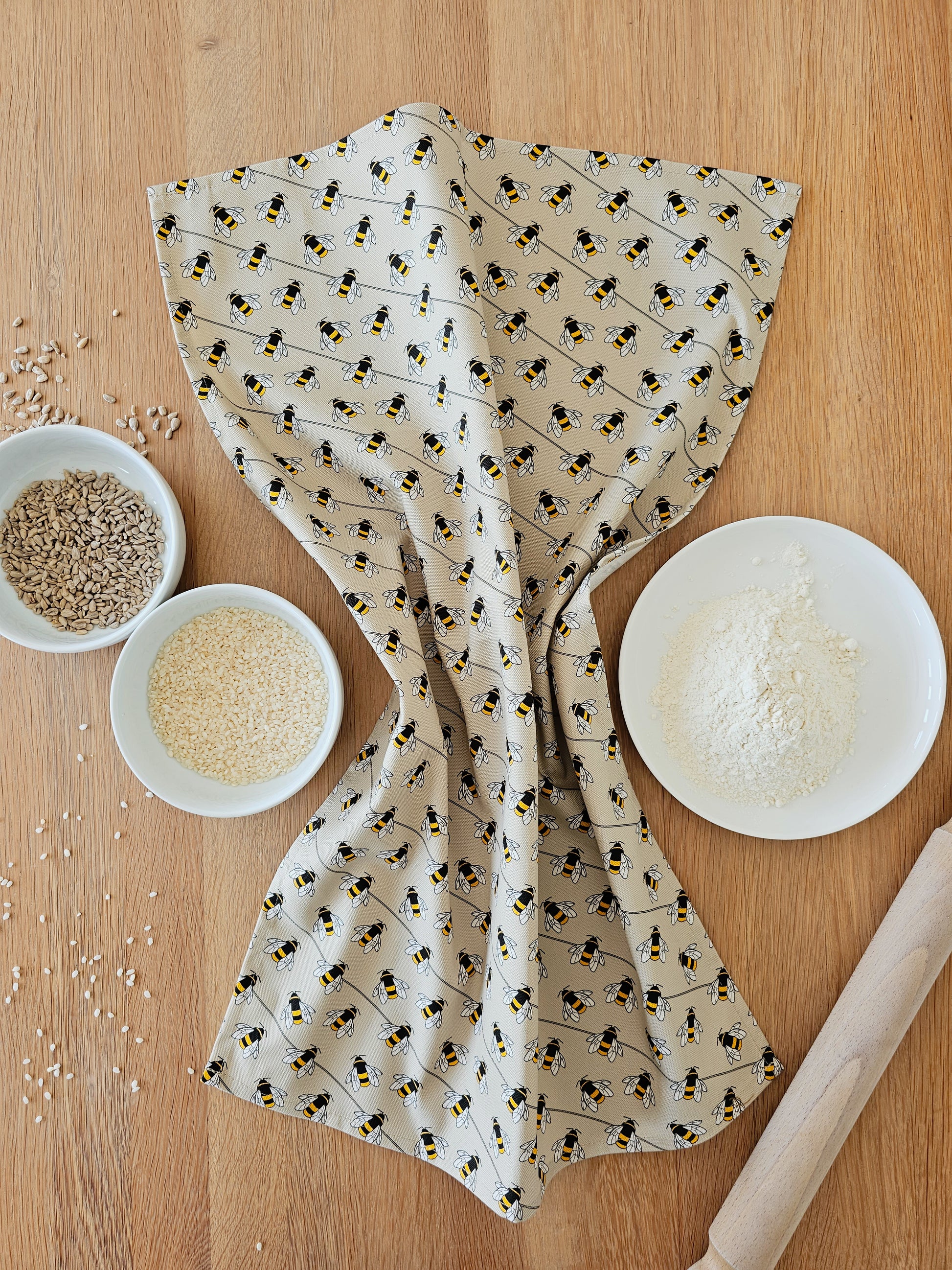 Organic cotton bee tea towel – designed by Hannah Issi and handmade in the UK. Stylish, sustainable kitchen linen. Shown on wooden table with cooking ingredients and rolling pin.