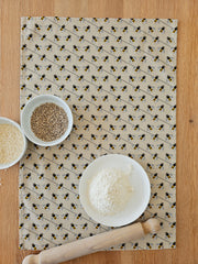 Organic cotton bee tea towel – designed by Hannah Issi and handmade in the UK. Stylish, sustainable kitchen linen. Shown laying on wooden table with cooking ingredients. 