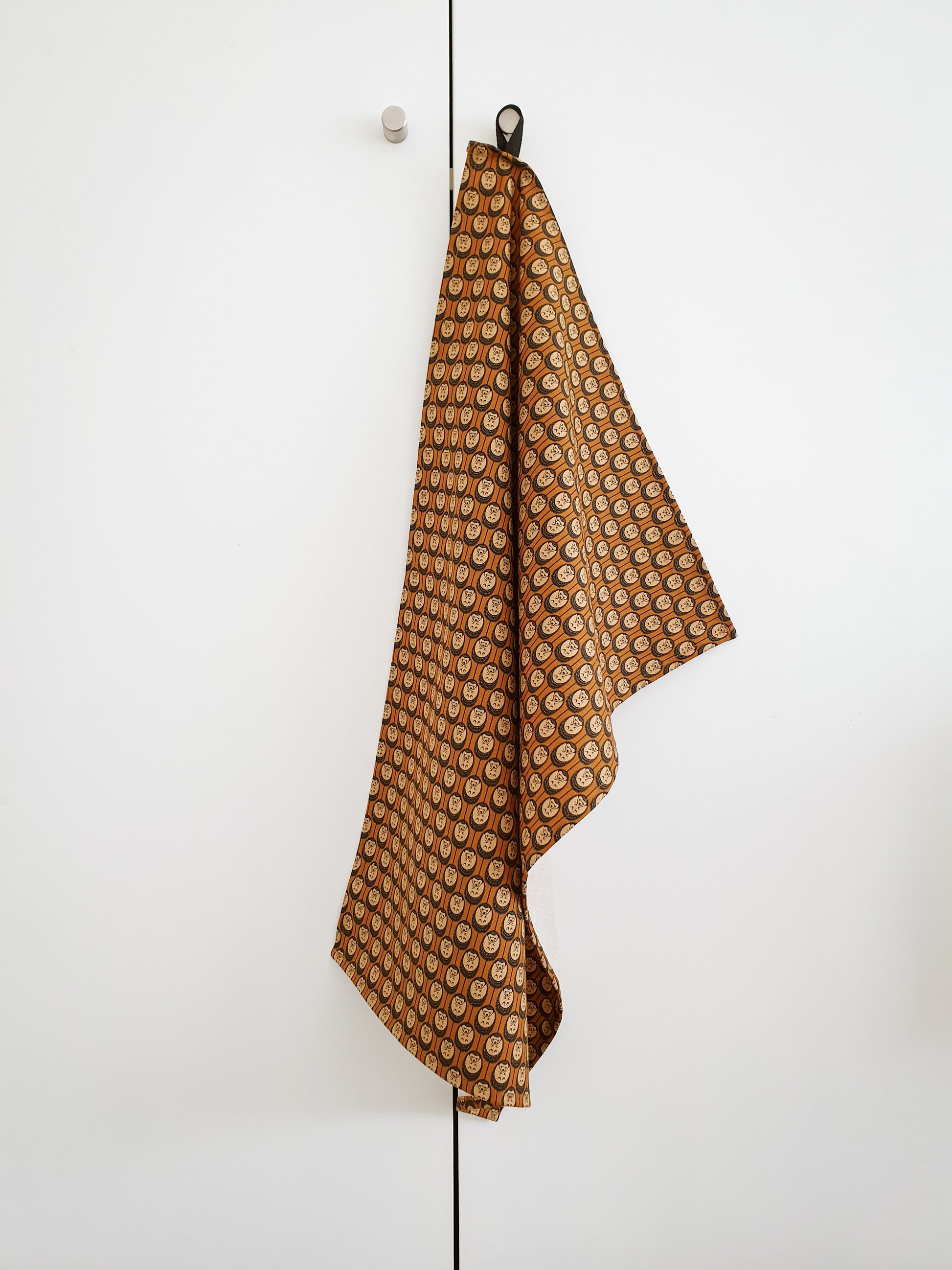 Organic cotton autumn hedgehog tea towel – burnt orange background, handmade in the UK by Hannah Issi. Stylish, sustainable linen shown hanging from door knob with white background. 