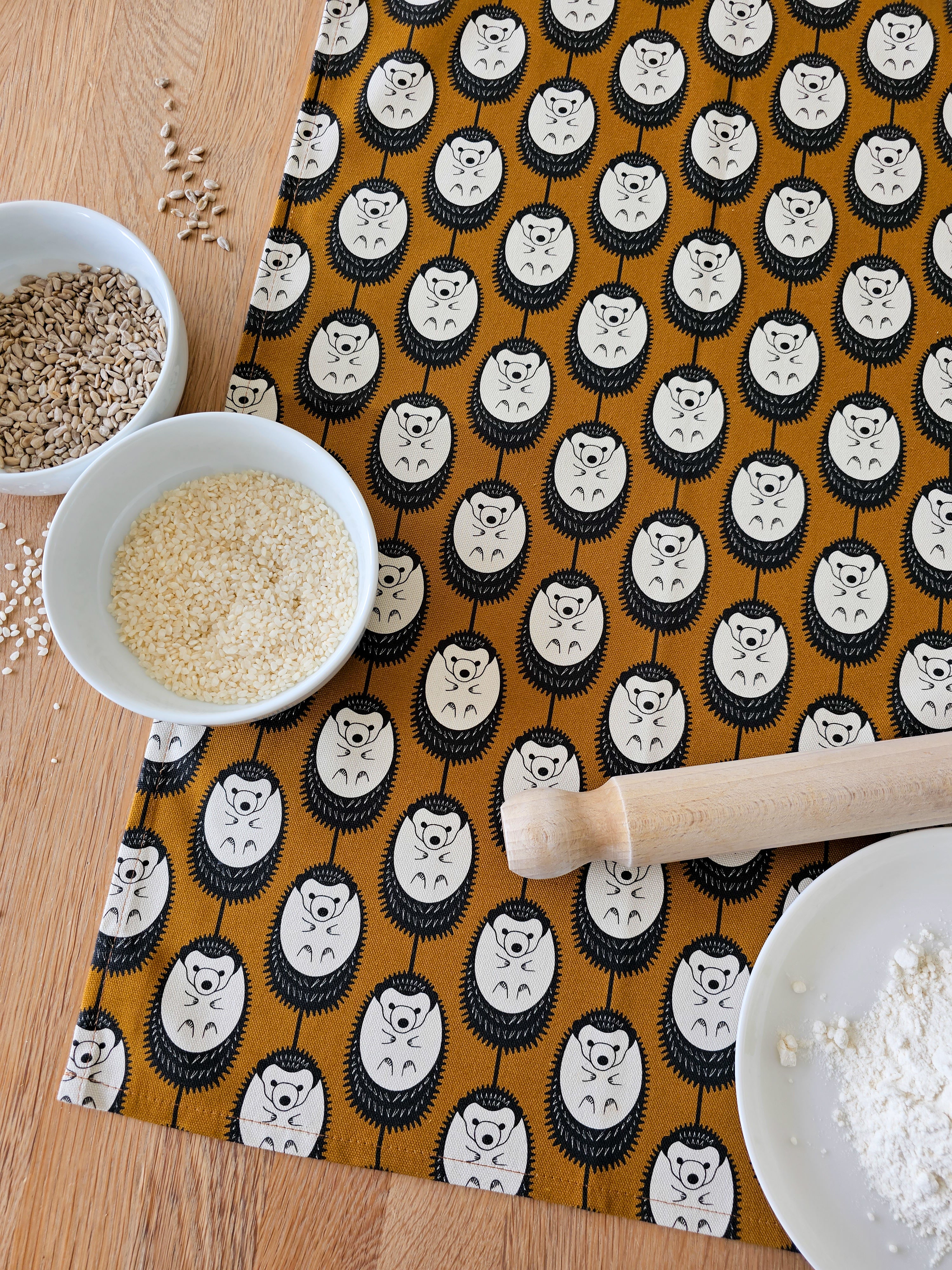 Organic cotton brown & white hedgehog tea towel – designed by Hannah Issi and handmade in the UK. Sustainable, modern and eco-friendly kitchen textile. Shown on wooden table with cooking ingredients and rolling pin. 