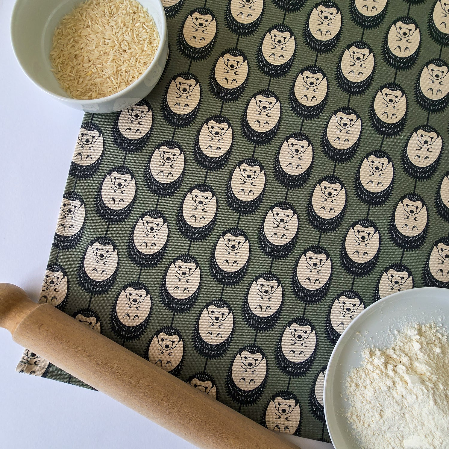 Organic cotton hedgehog tea towel in new green & white colour – designed by Hannah Issi and handmade in the UK. Sustainable, modern and eco-friendly kitchen textile. Lifestyle image showing close up of tea towel on white background with cooking ingredients and rolling pin.