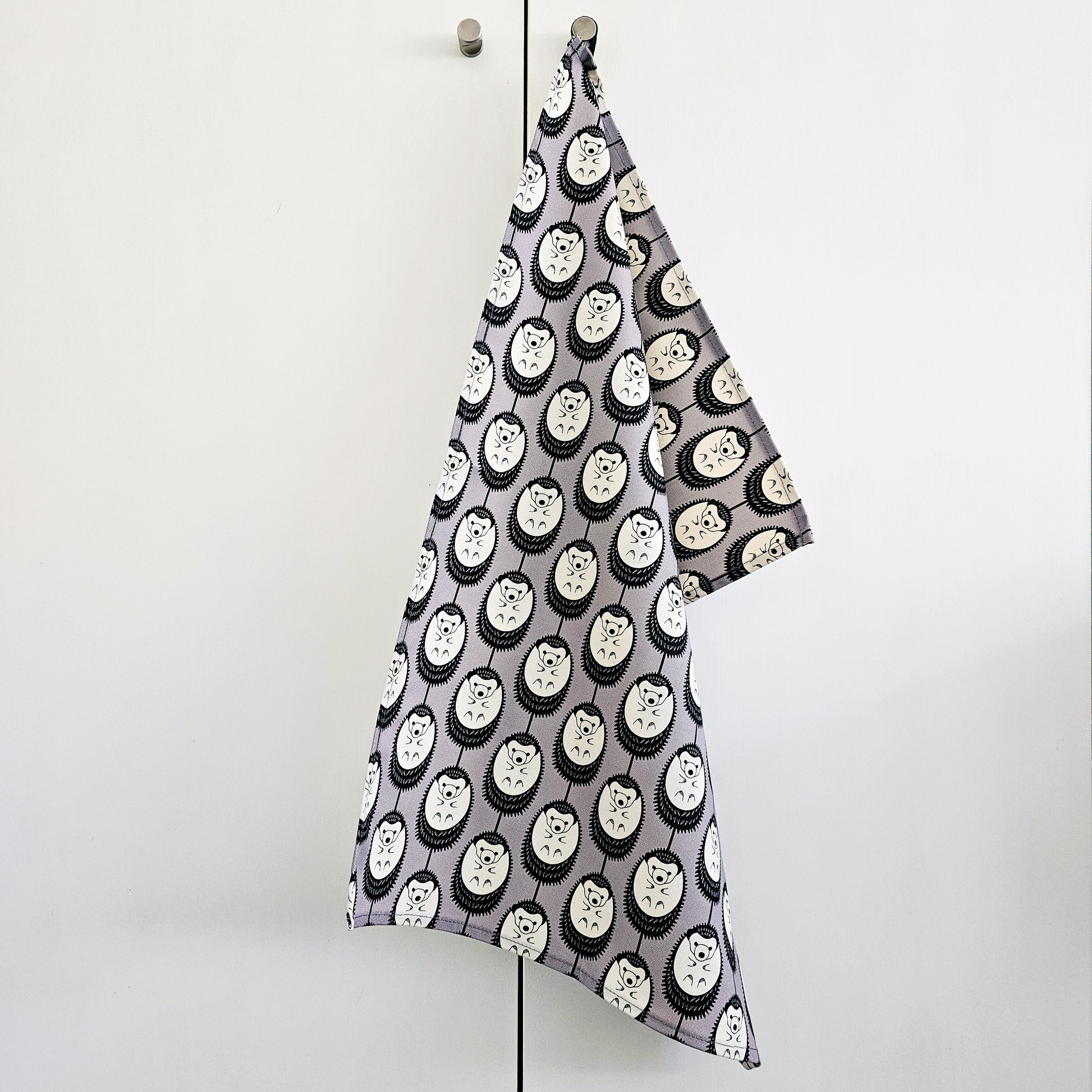 Organic cotton grey and white hedgehog tea towel – designed by Hannah Issi and handmade in the UK. Stylish, sustainable kitchen linen- shown hanging on white door from door knob.