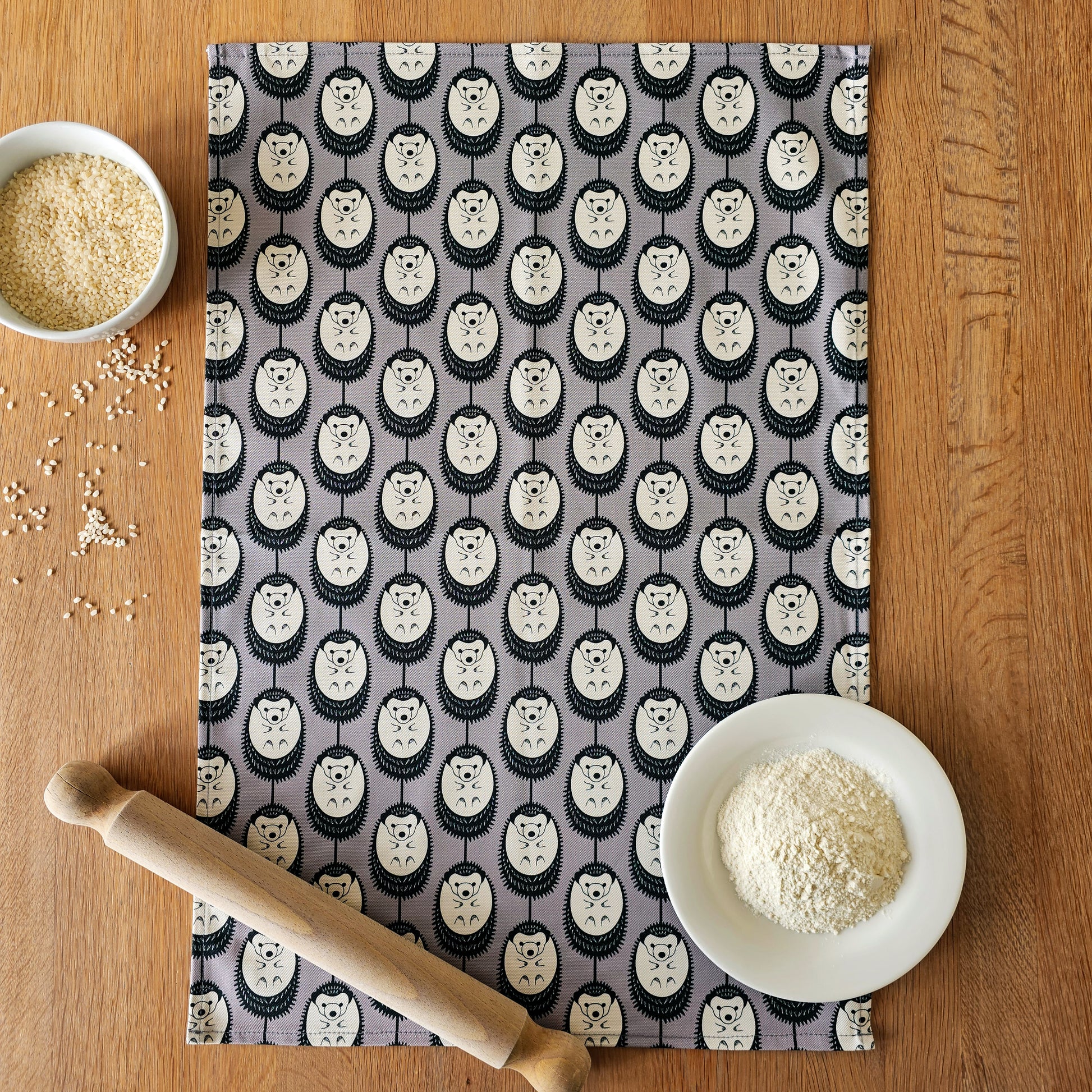 Organic cotton grey and white hedgehog tea towel – designed by Hannah Issi and handmade in the UK. Stylish, sustainable kitchen linen- laying on wooden tabletop with wooden rolling pin and cooking ingredients. 