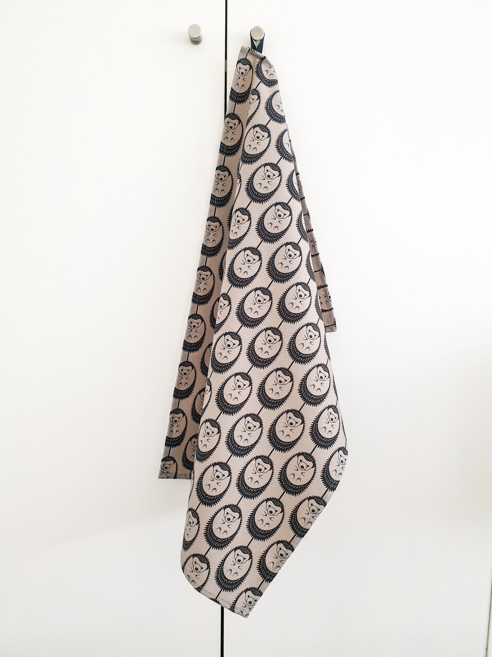 Organic cotton grey hedgehog tea towel – designed by Hannah Issi and handmade in the UK. Stylish, sustainable kitchen linen shown hanging on door knob with white background.