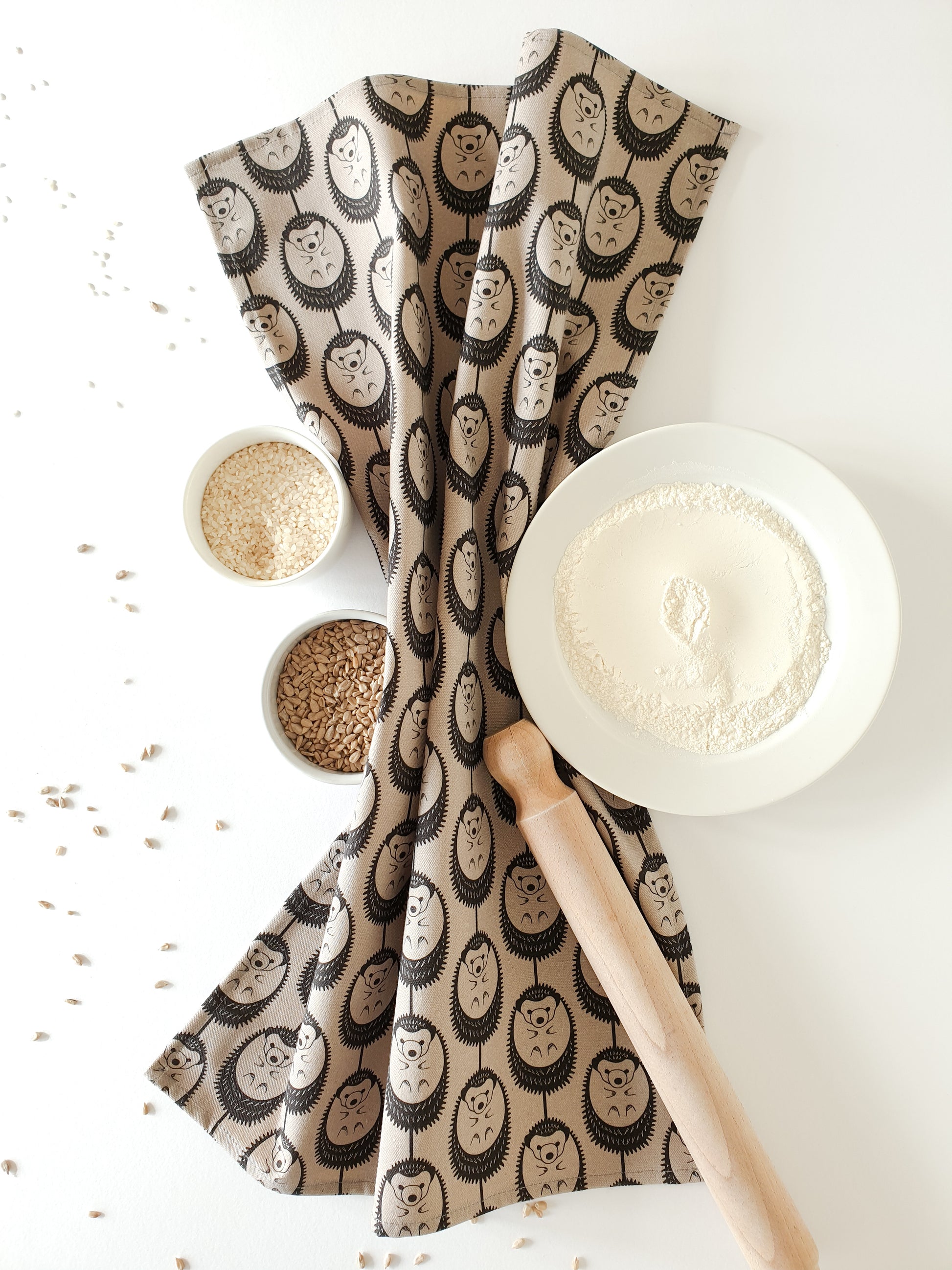 Organic cotton grey hedgehog tea towel – designed by Hannah Issi and handmade in the UK. Stylish, sustainable kitchen linen shown on white background with rolling pin and cooking ingredients.