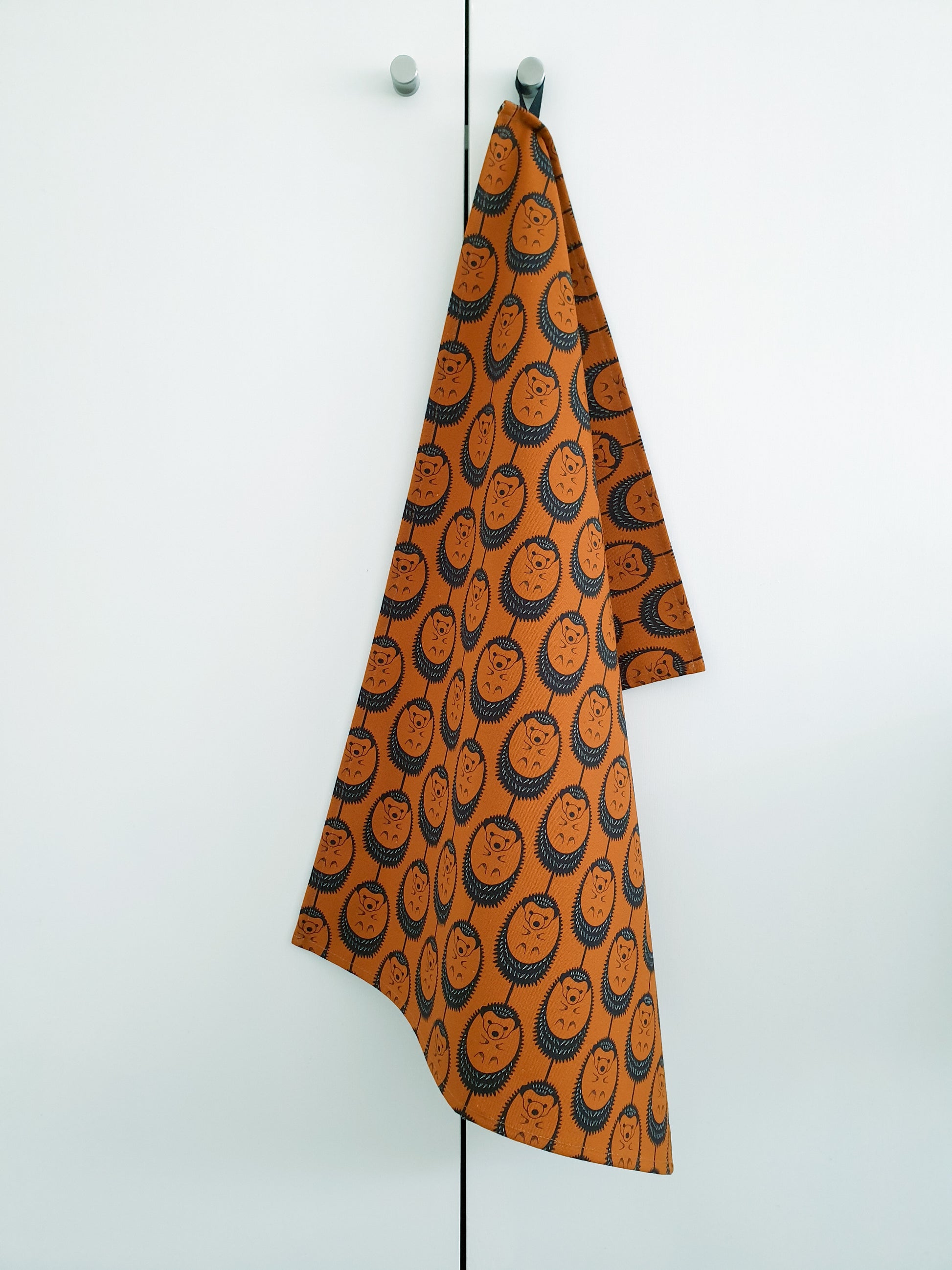 Organic cotton terracotta hedgehog tea towel – designed by Hannah Issi and handmade in the UK. Stylish, sustainable kitchen linen - shown hanging from door knob with white background.
