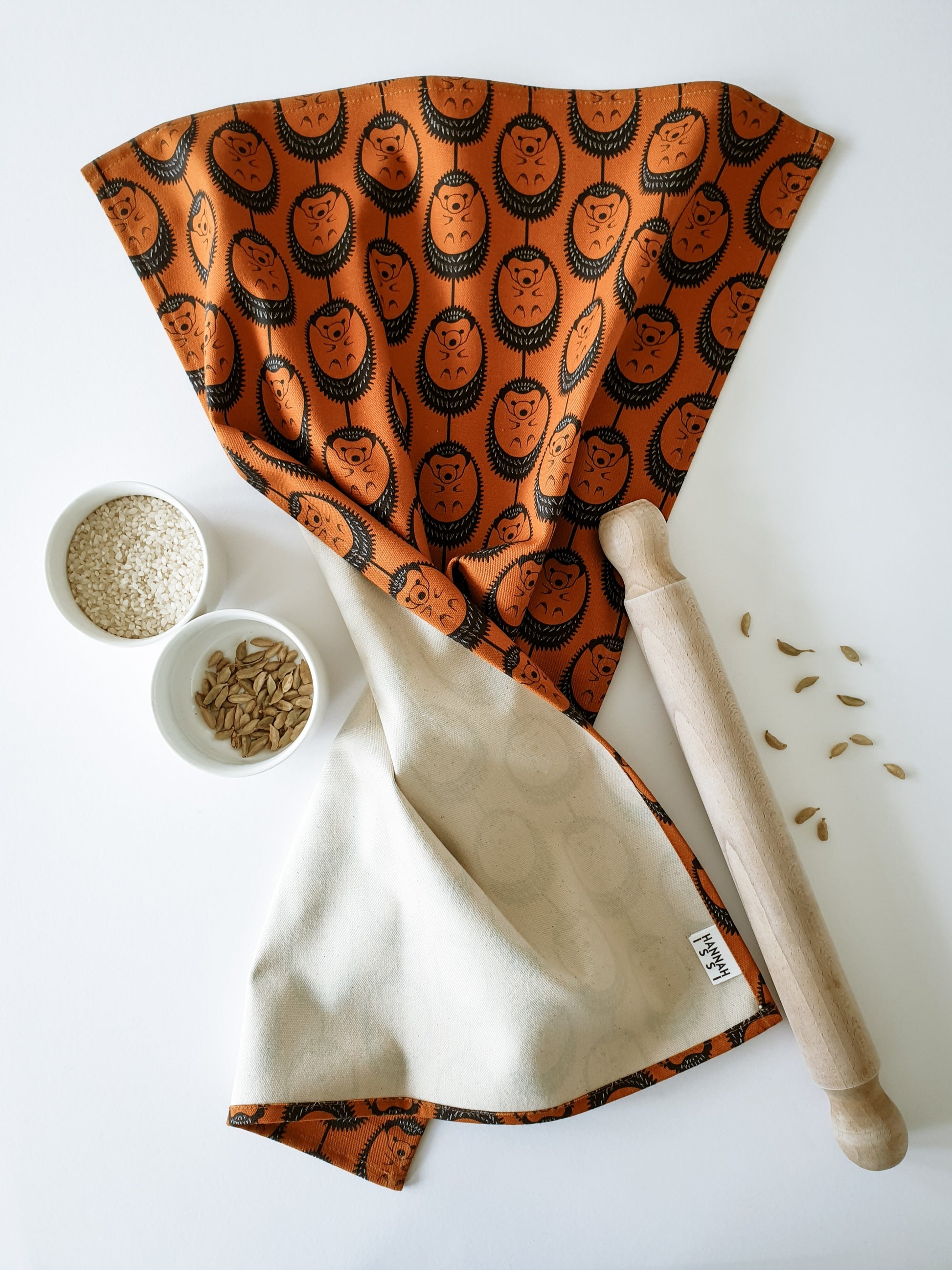 Organic cotton terracotta hedgehog tea towel – designed by Hannah Issi and handmade in the UK. Stylish, sustainable kitchen linen - shown laying on a white background and cooking ingredients. 