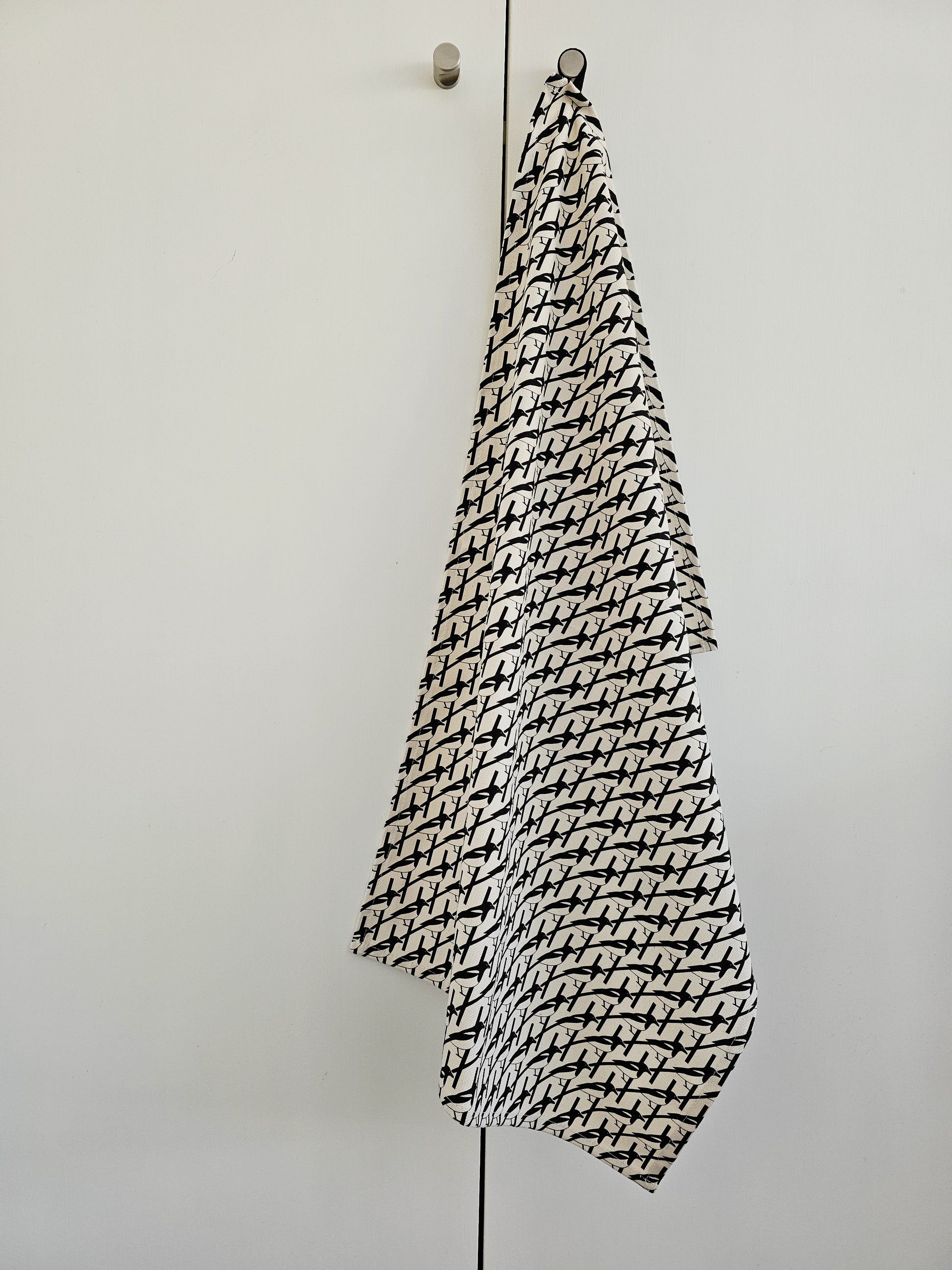 Organic cotton black and white magpie tea towel – designed by Hannah Issi and handmade in the UK. Stylish, sustainable kitchen linen shown hanging from door knob with white background.