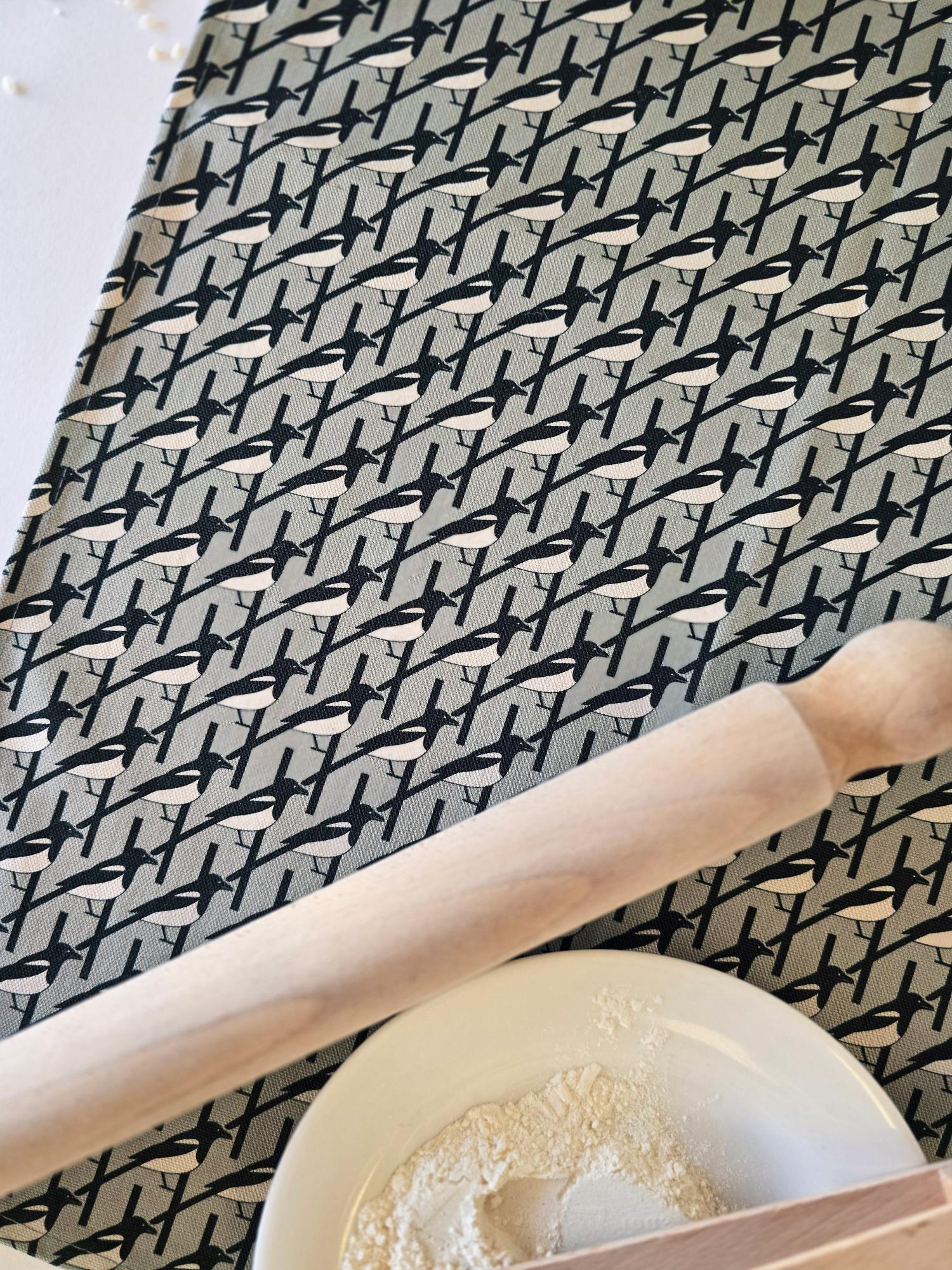 Organic cotton grey magpie tea towel – designed by Hannah Issi and handmade in the UK. Stylish, sustainable kitchen linen. Close- up image shows Magpie bird pattern on tea towel with white background and cooking ingredients.