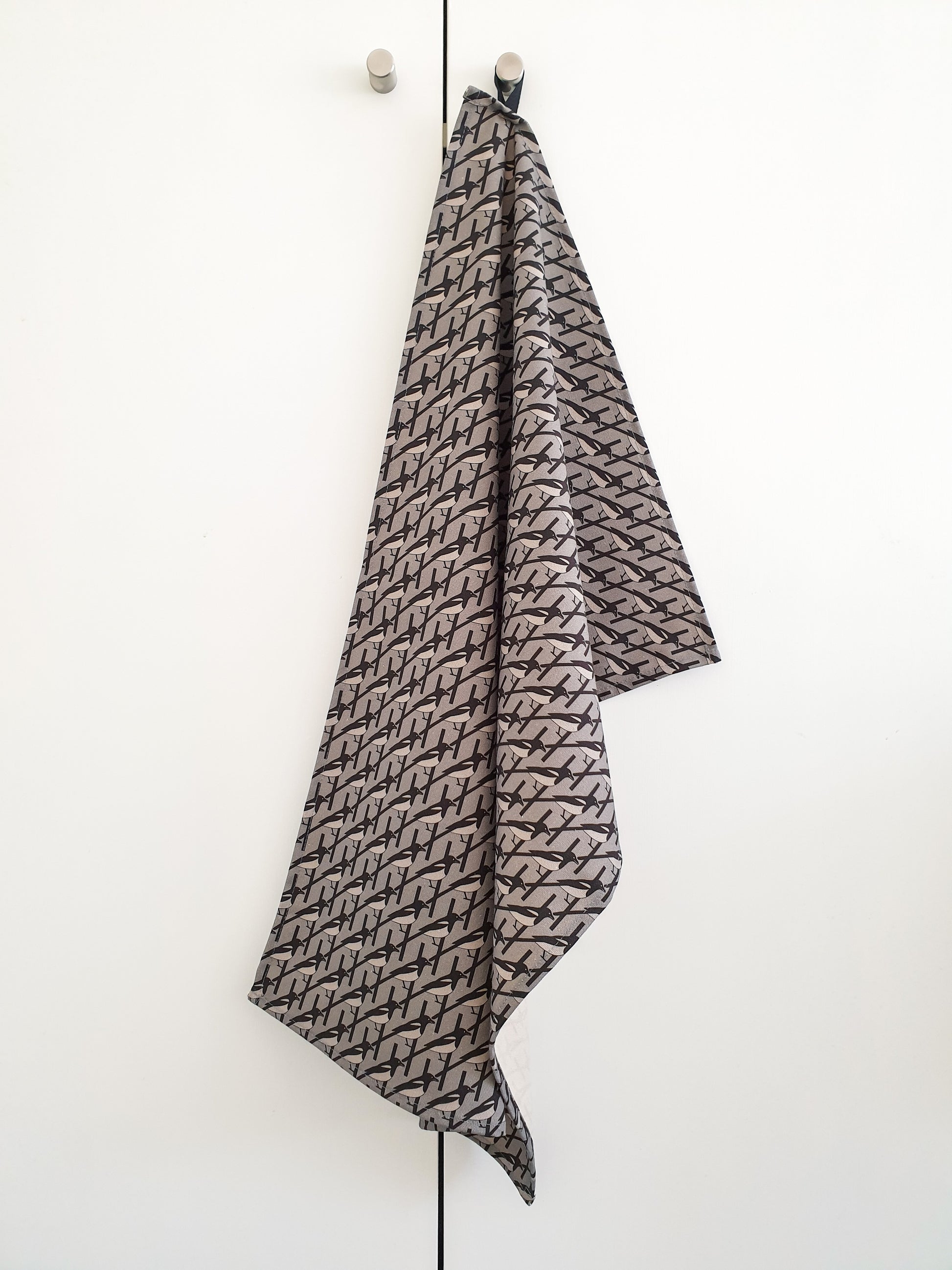 Organic cotton grey magpie tea towel – designed by Hannah Issi and handmade in the UK. Stylish, sustainable kitchen linen shown hanging on door knob with white background.
