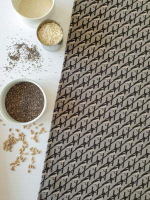 Organic cotton grey magpie tea towel – designed by Hannah Issi and handmade in the UK. Stylish, sustainable kitchen linen. Image shows tea towel with white background and cooking ingredients.