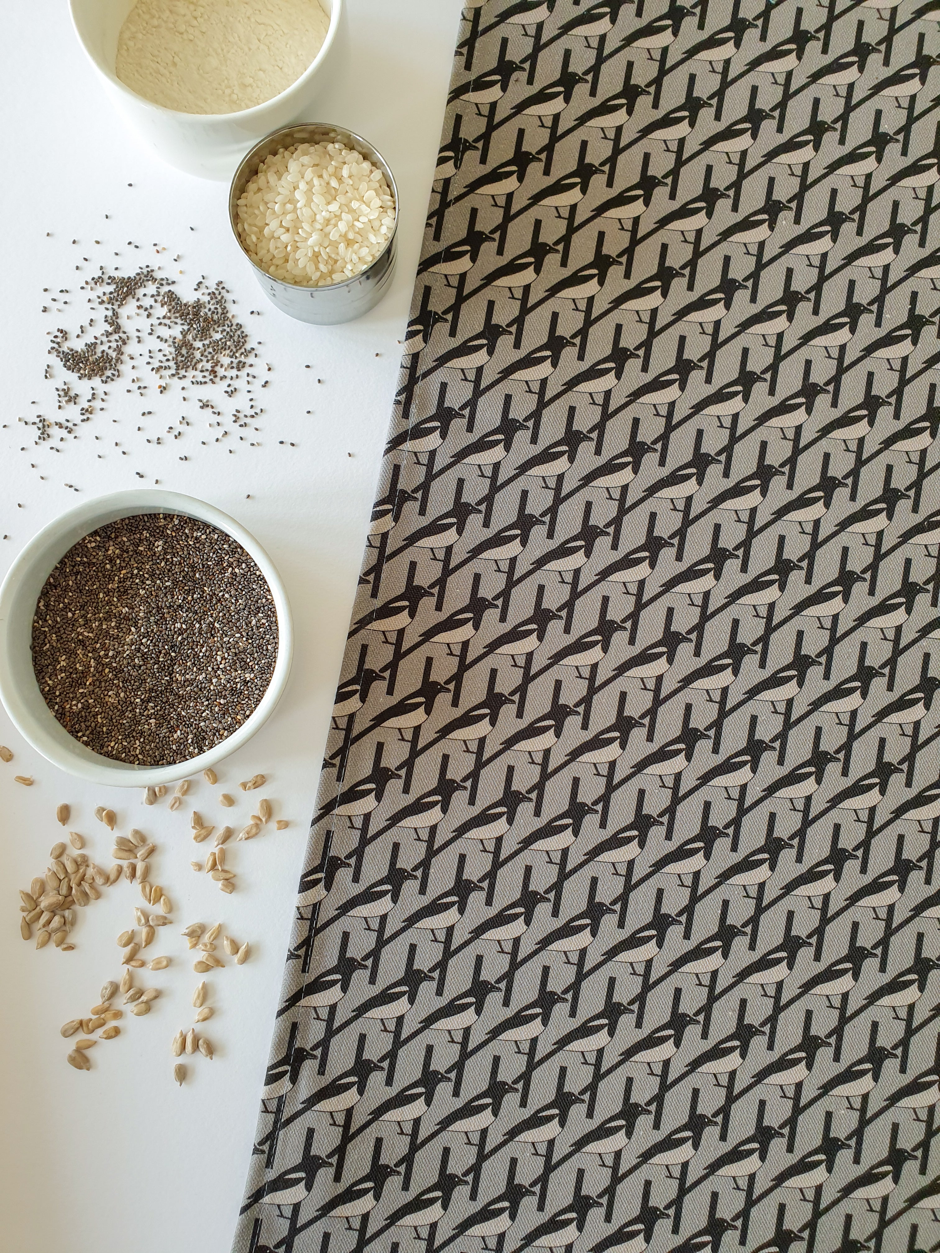 Organic cotton grey magpie tea towel – designed by Hannah Issi and handmade in the UK. Stylish, sustainable kitchen linen. Image shows tea towel with white background and cooking ingredients.