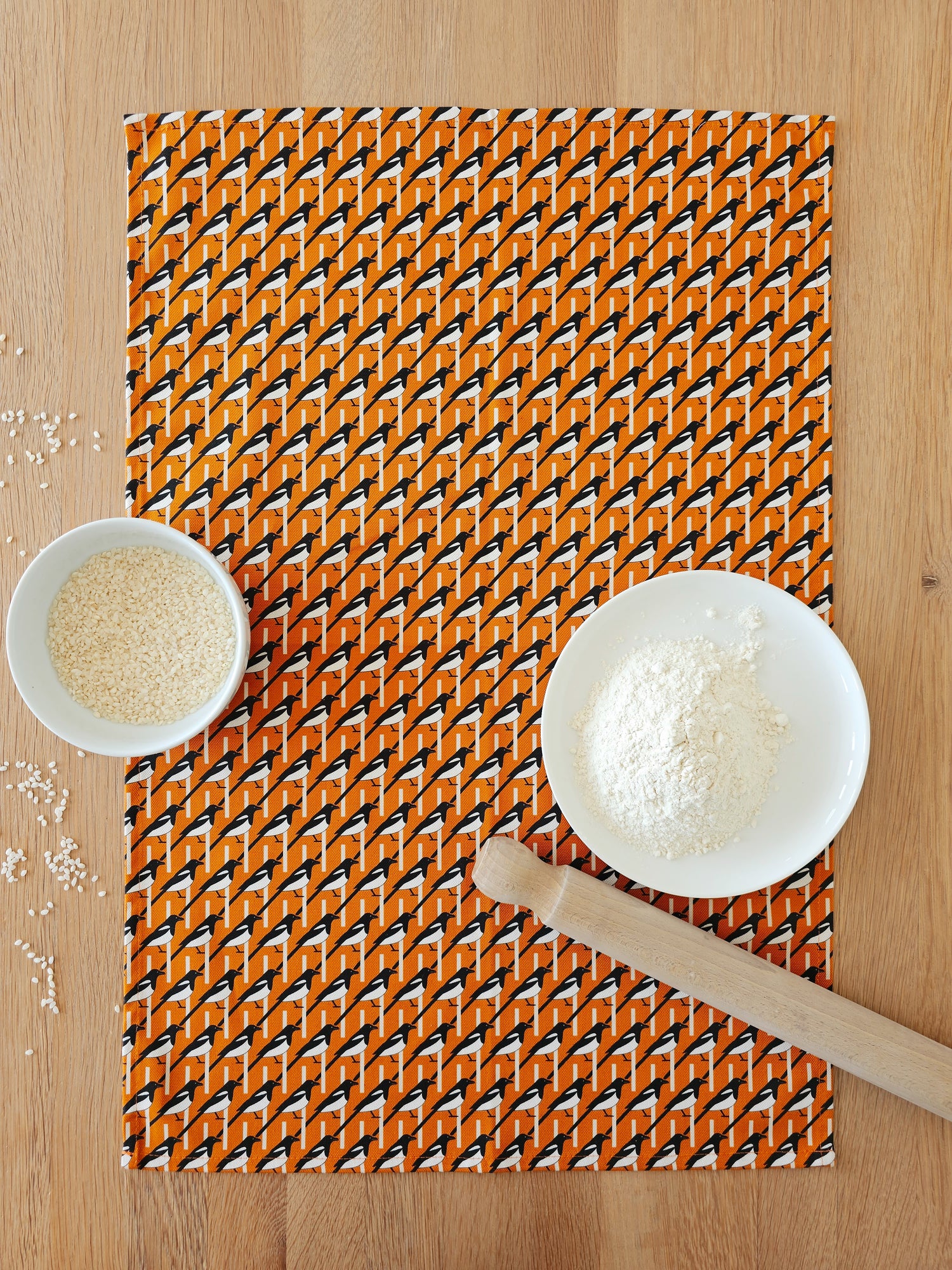 Organic cotton orange magpie tea towel – designed by Hannah Issi and handmade in the UK. Stylish, sustainable kitchen linen. Lifestyle image on wooden tabletop with cooking ingredients. 