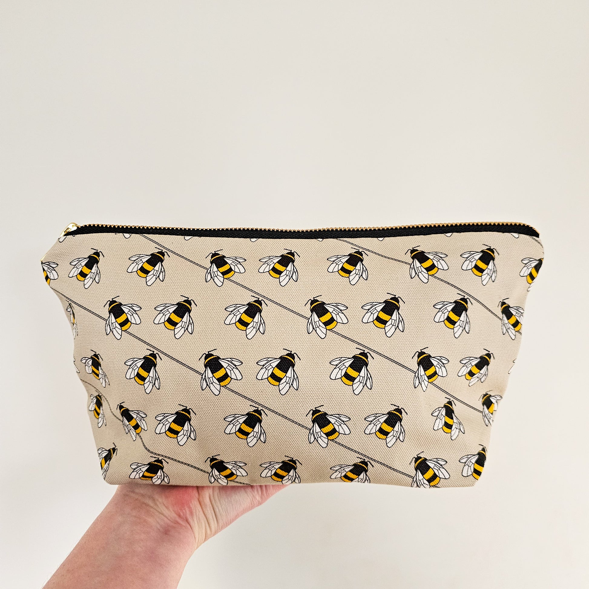 Organic cotton bee zip bag – handmade in the UK by Hannah Issi. Taupe bee print, versatile and sustainable accessory. Hand holding bag with neutral background.